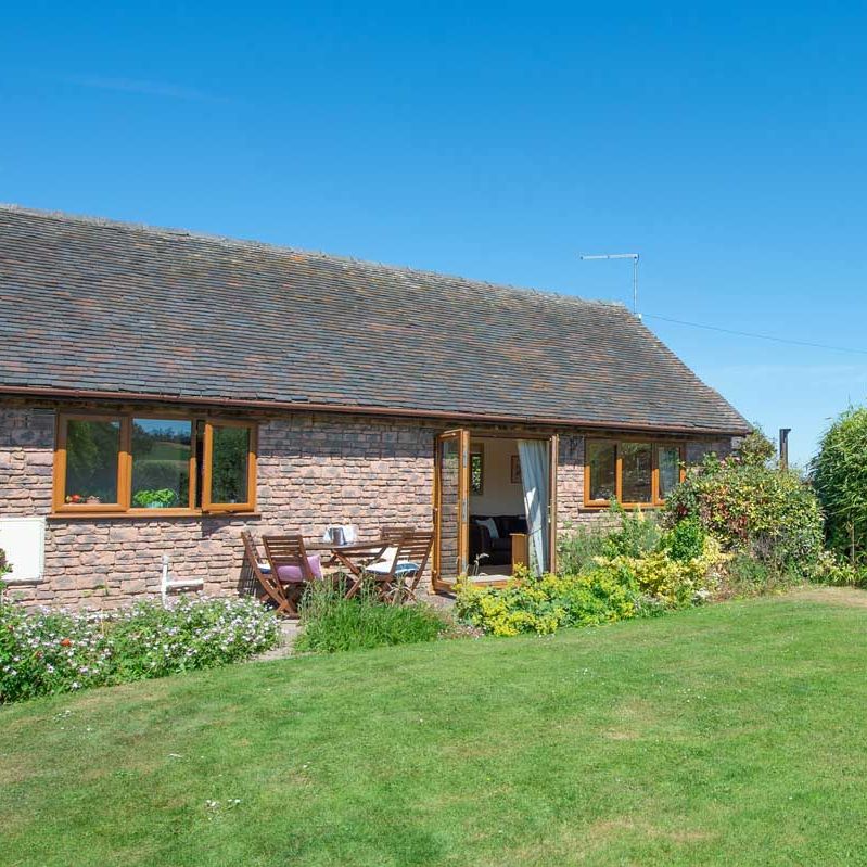 Single-story brick cottage with wooden windows, surrounded by a green lawn and garden under a clear blue sky.