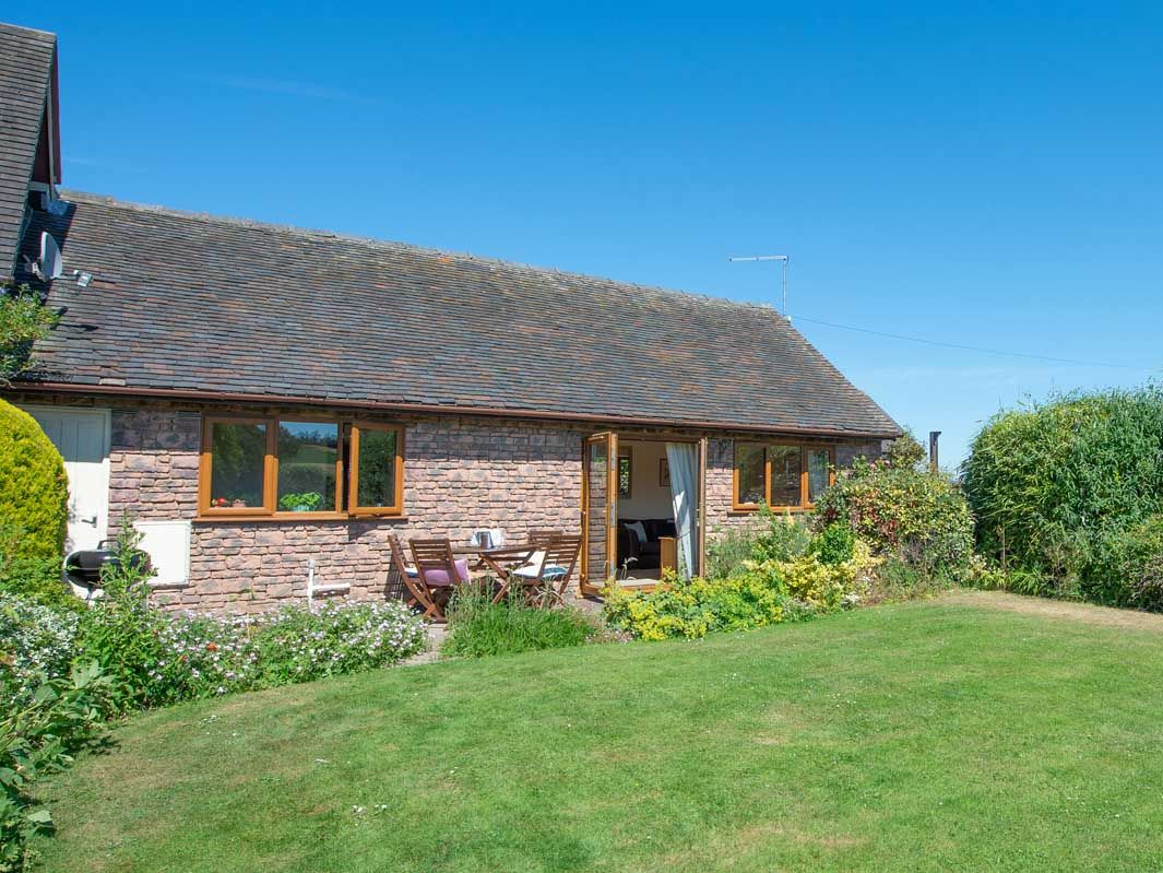 Single-story brick cottage with wooden windows, surrounded by a green lawn and garden under a clear blue sky.