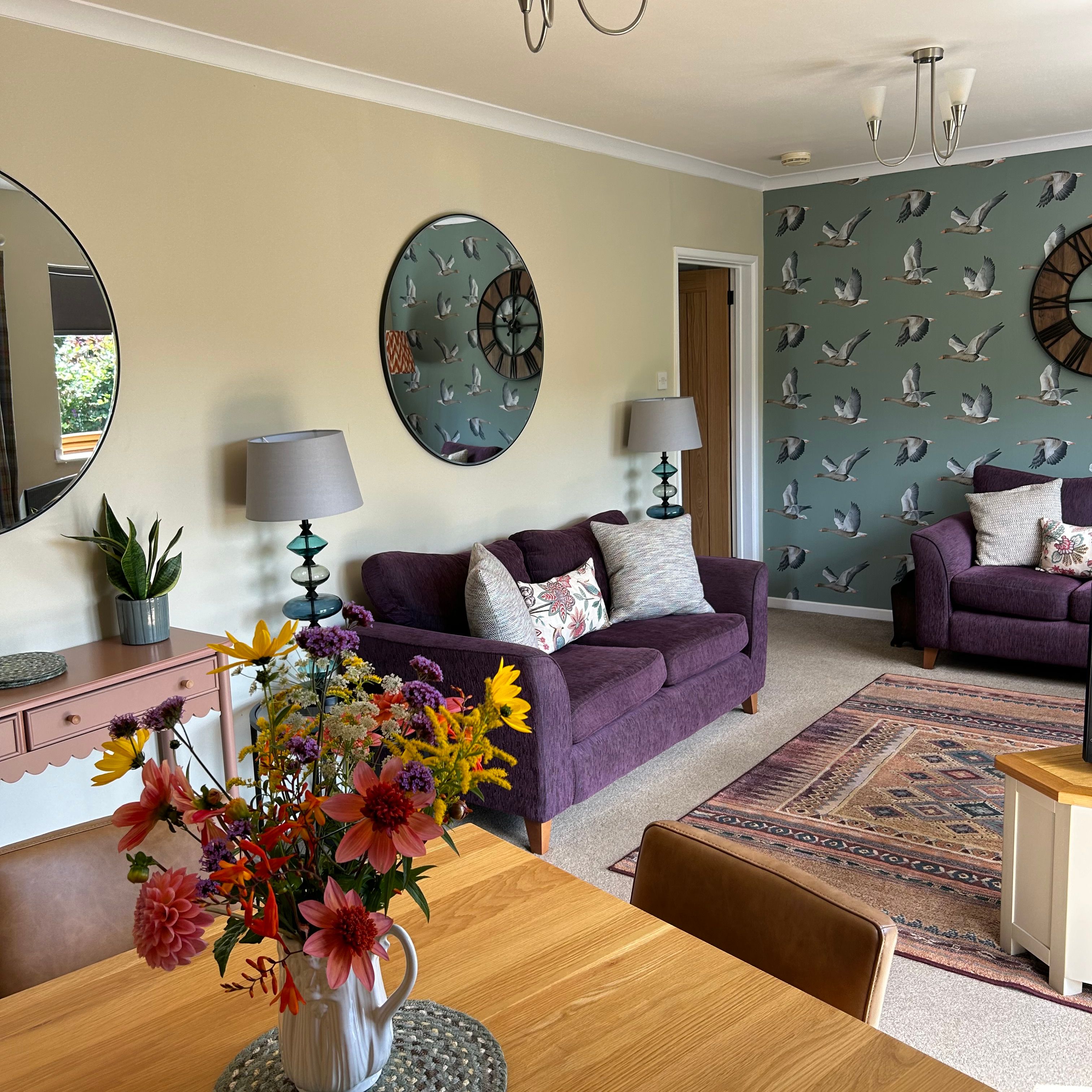 Bright living room with purple sofas, bird wallpaper, and a vase of colorful flowers on a wooden table.