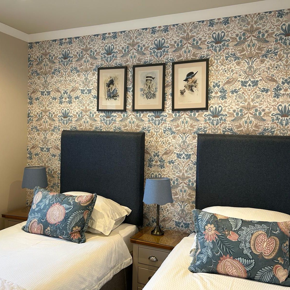 Twin beds with patterned pillows, blue lamps, and floral wallpaper in a bedroom.