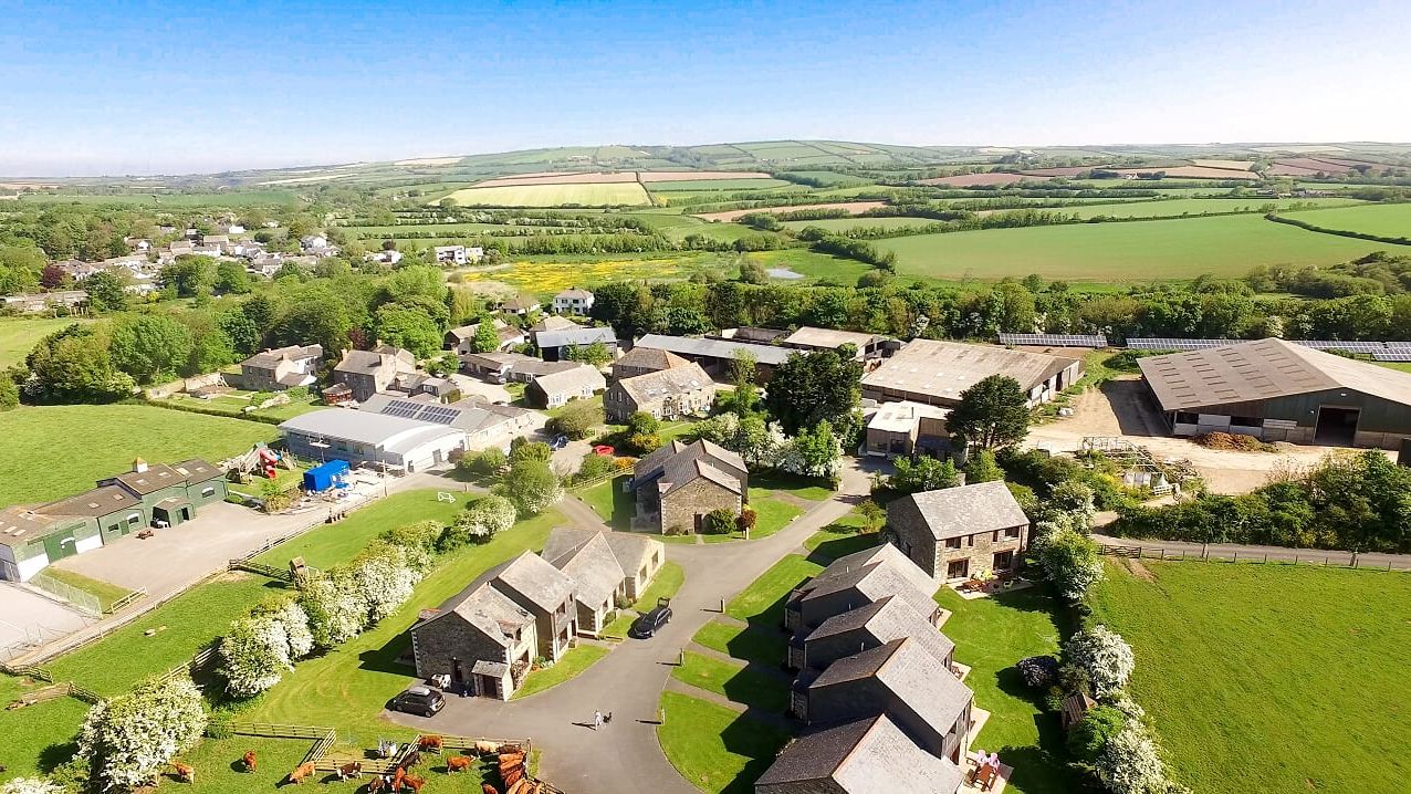 Aerial view of a rural village with stone houses, barns, green fields, and farmland under a bright blue sky.