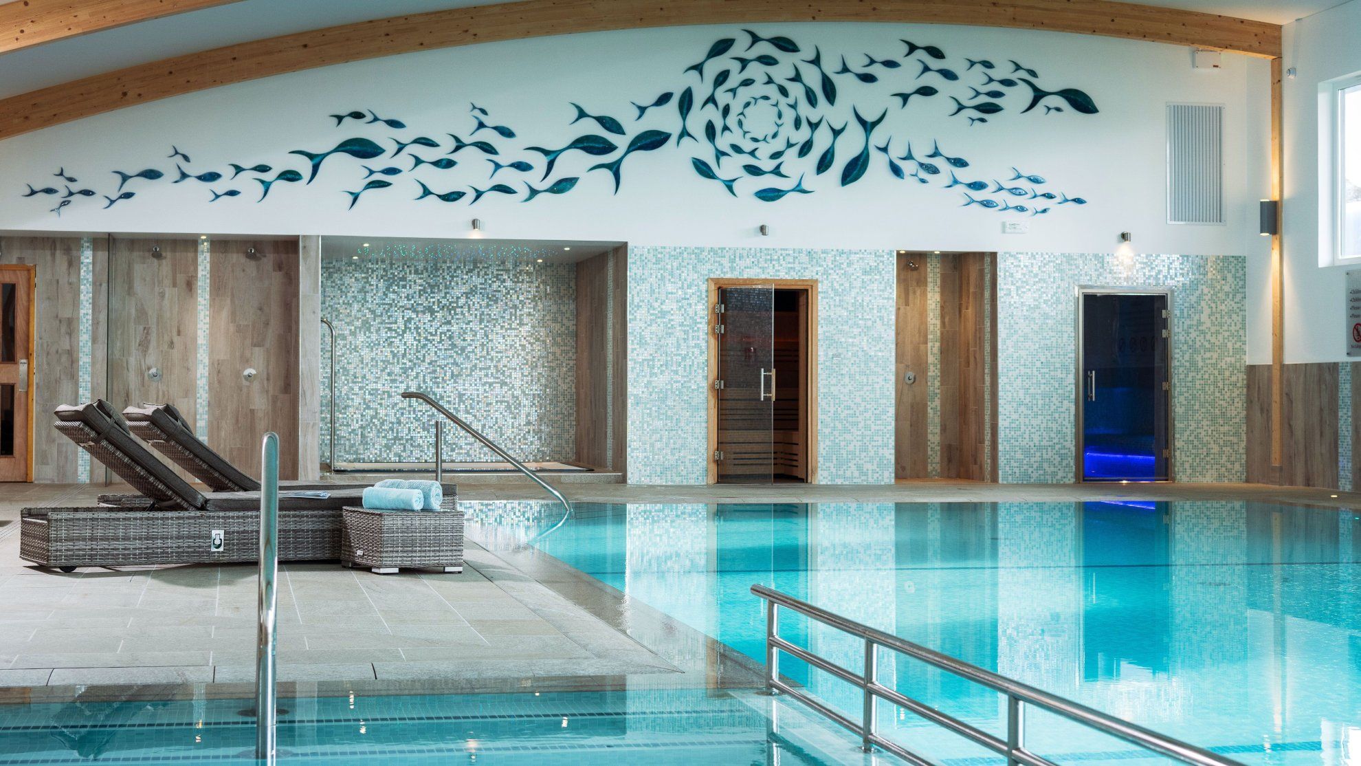 Indoor swimming pool with lounge chairs, a mosaic tile wall, and a fish mural above