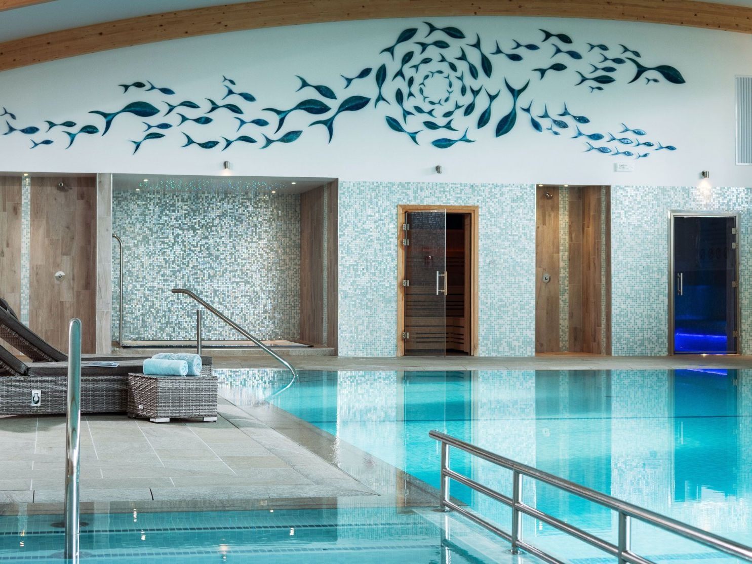 Indoor swimming pool with lounge chairs, a mosaic tile wall, and a fish mural above