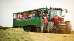 A red tractor pulling a green wagon filled with people, including children and adults, enjoying a ride through a grassy field. The people are smiling and waving.