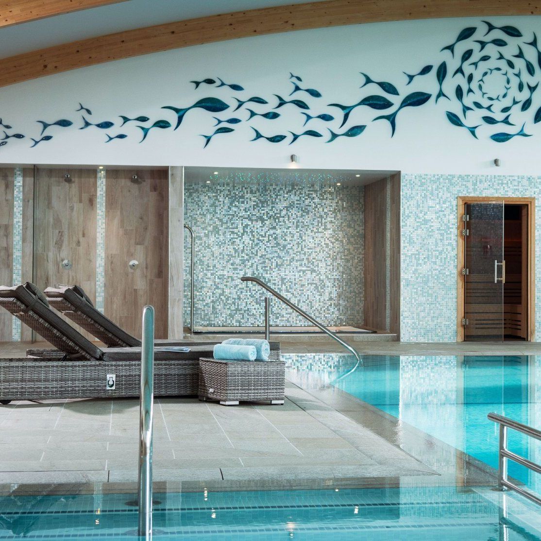 Modern indoor pool area with lounge chairs, wooden doors, mosaic tiled walls, and a fish mural on the upper wall.
