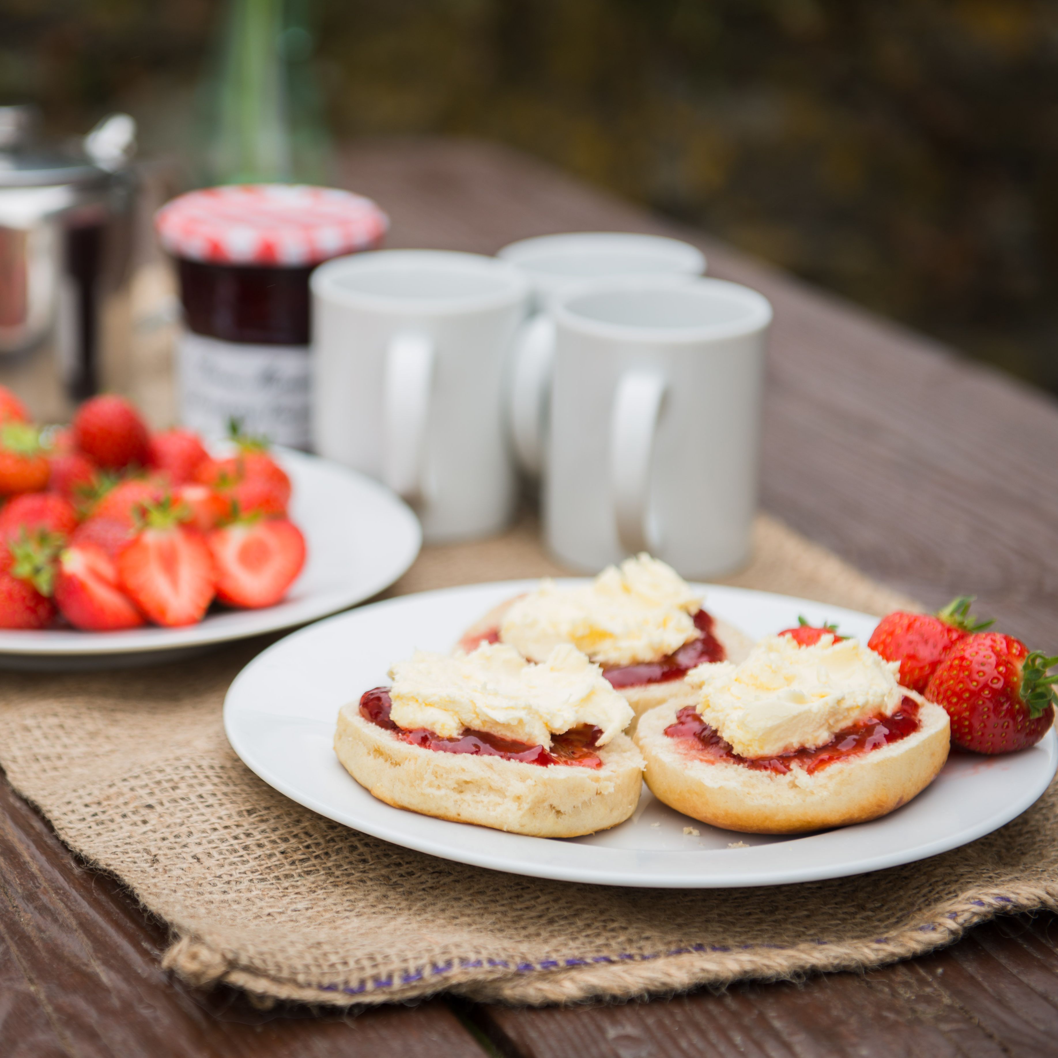 A rustic outdoor table set with plates of strawberries, scones topped with clotted cream and jam, three white mugs, a jar of jam, and a metal teapot.