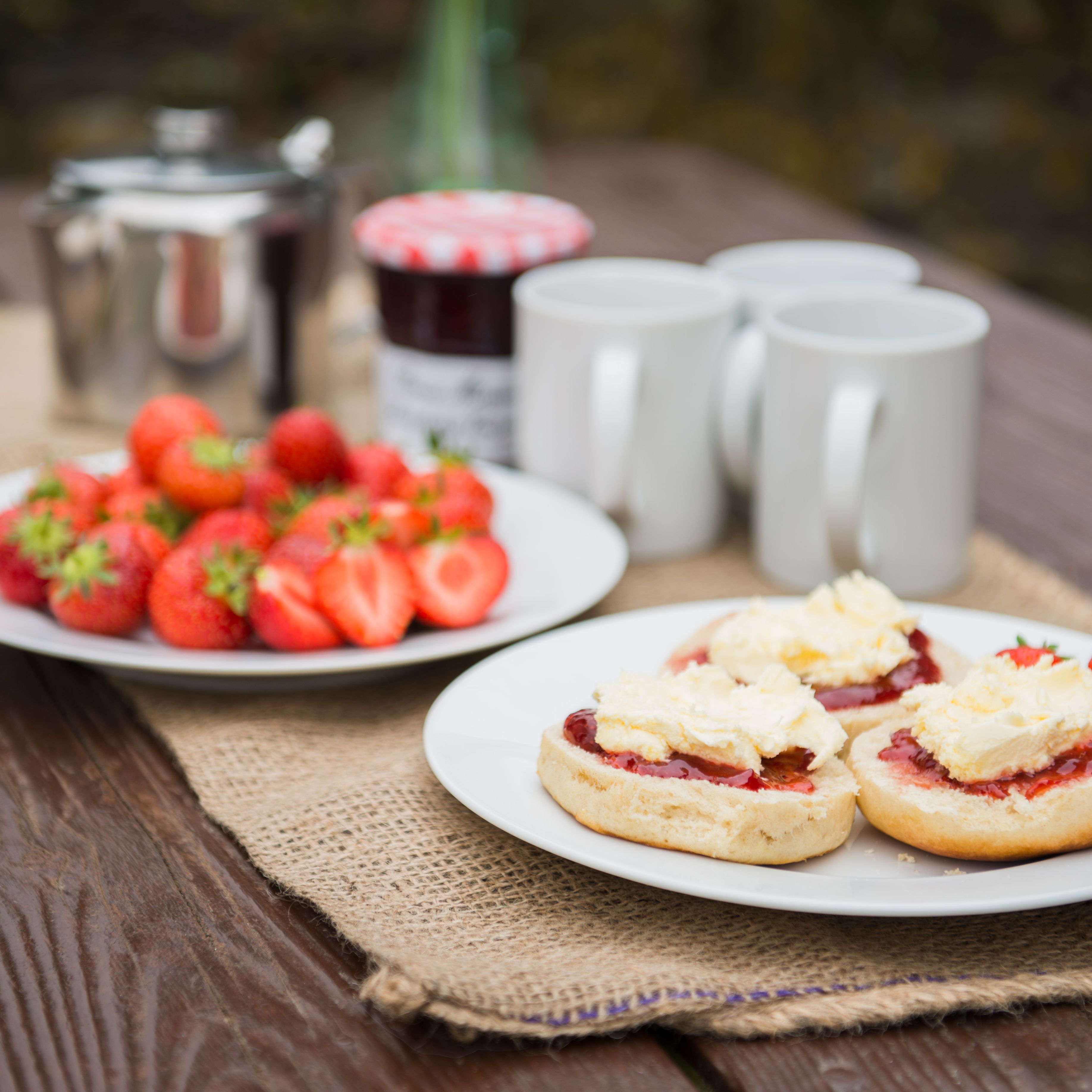 A rustic outdoor table set with plates of strawberries, scones topped with clotted cream and jam, three white mugs, a jar of jam, and a metal teapot.