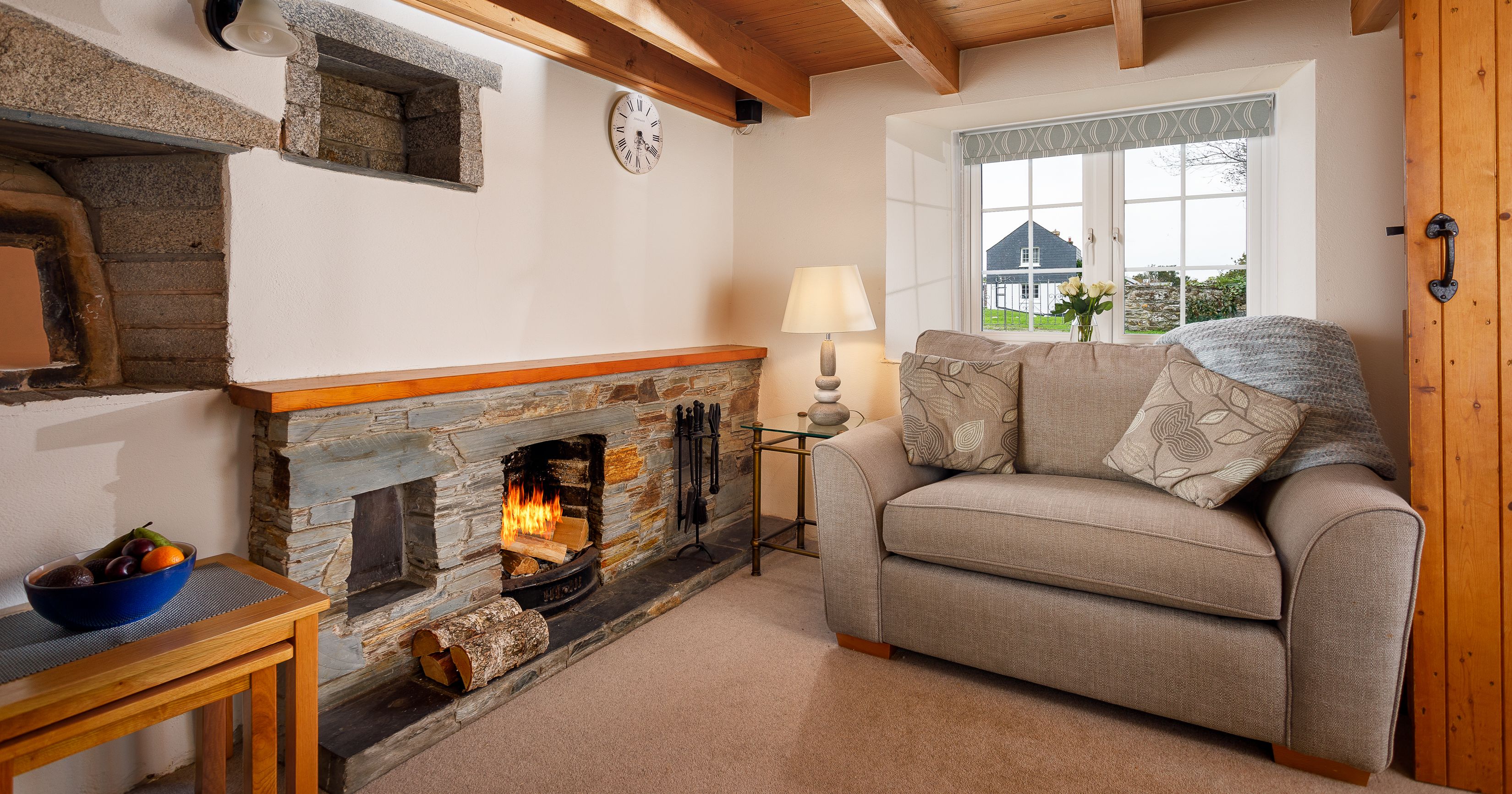 Cozy living room with stone fireplace, beige sofa, wooden beams, and window view.