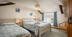 Cozy bedroom with a double and a single bed, floral bedding, wooden beams, and a window with blue curtains.