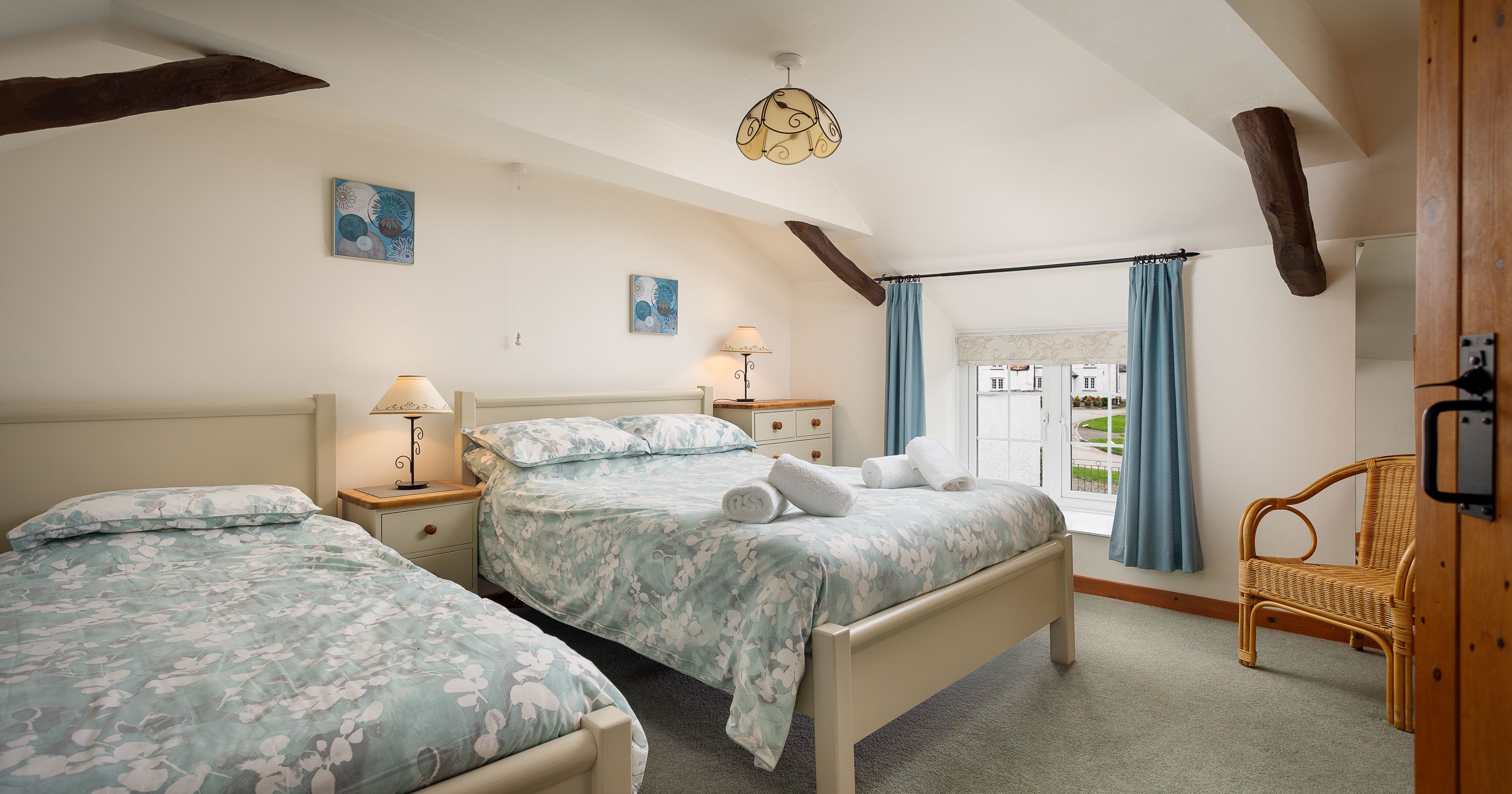 Cozy bedroom with a double and a single bed, floral bedding, wooden beams, and a window with blue curtains.