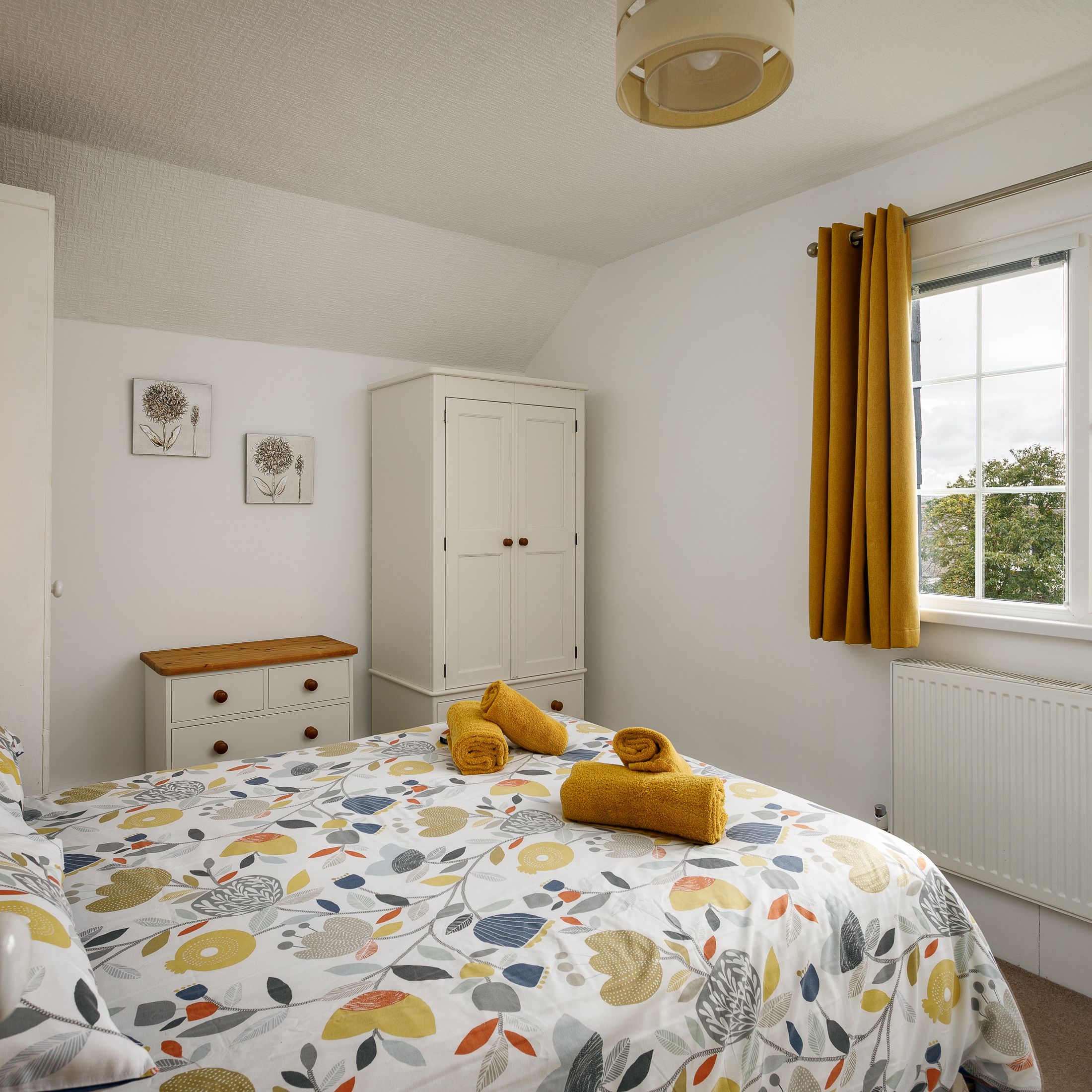 Bright, cozy bedroom with floral bedding, yellow towels on the bed, white furniture, and a window with yellow curtains.