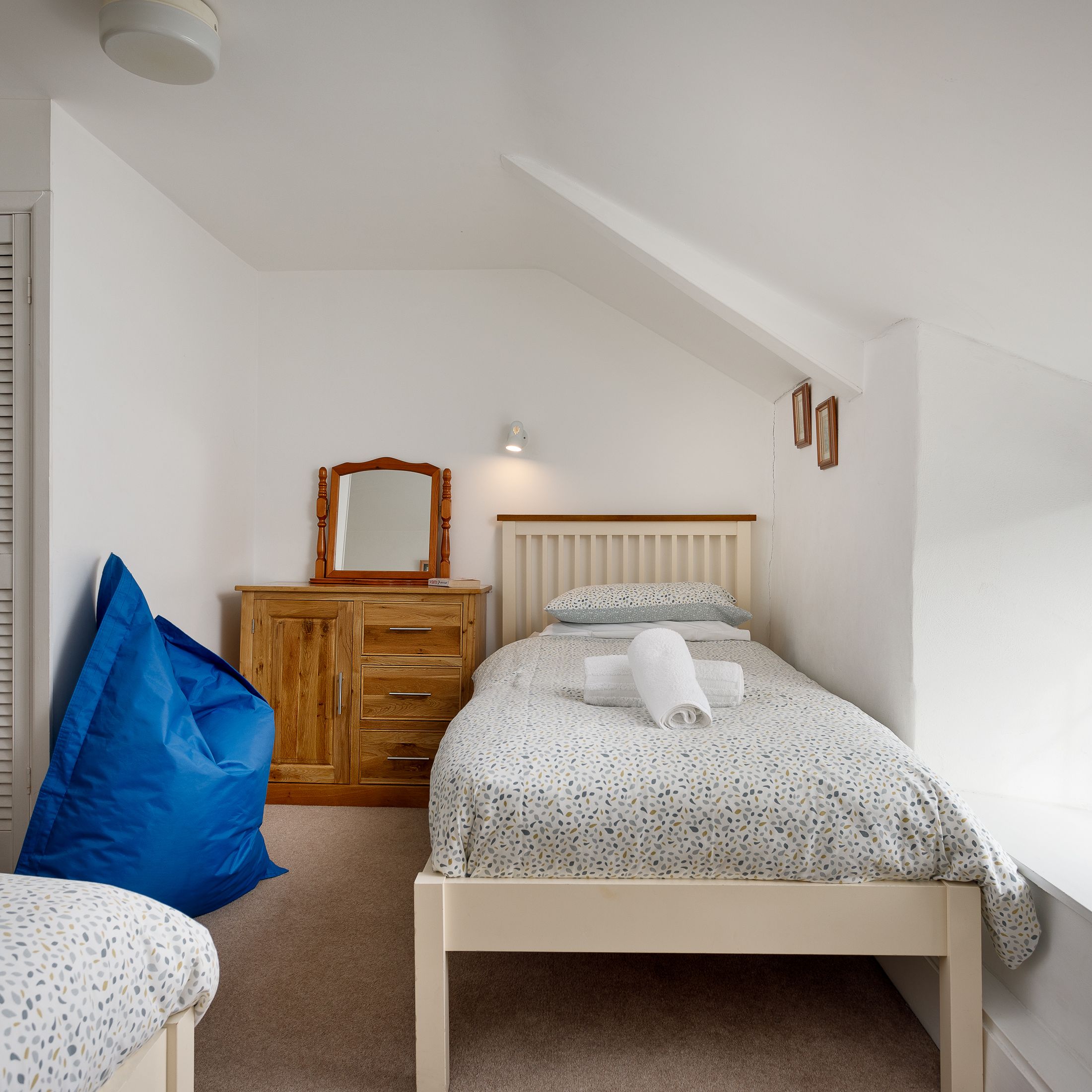 Small cozy bedroom with twin beds, a wooden dresser with mirror, blue bean bag, and large window with natural light.