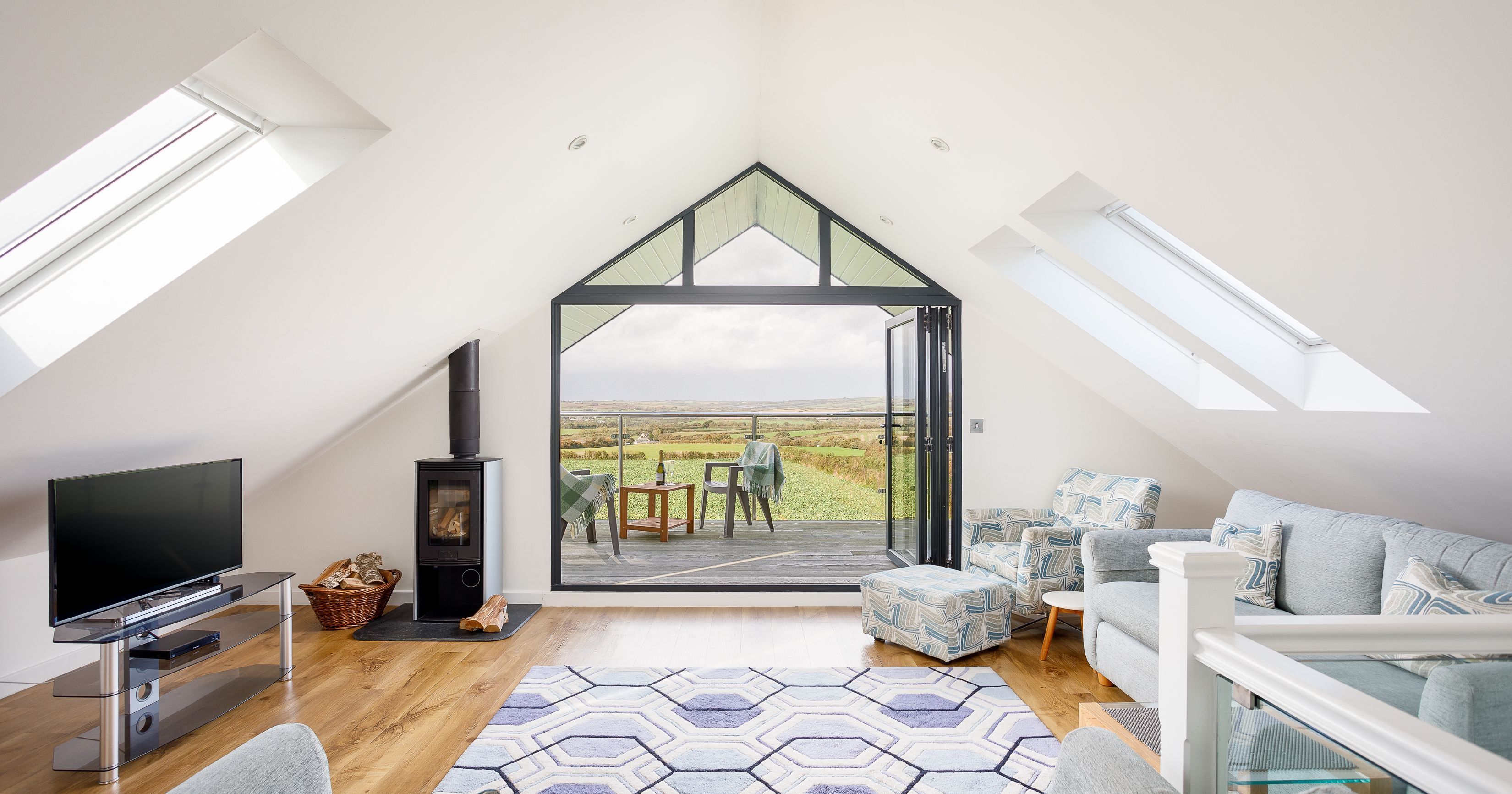 Modern attic living room with large glass doors opening to a balcony overlooking green fields.