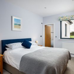 Modern double bedroom with a blue headboard, light bedding, wooden furniture, and a window with countryside views