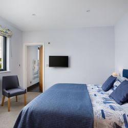 Modern bedroom with blue bedding, wall-mounted TV, large window, and en-suite bathroom.