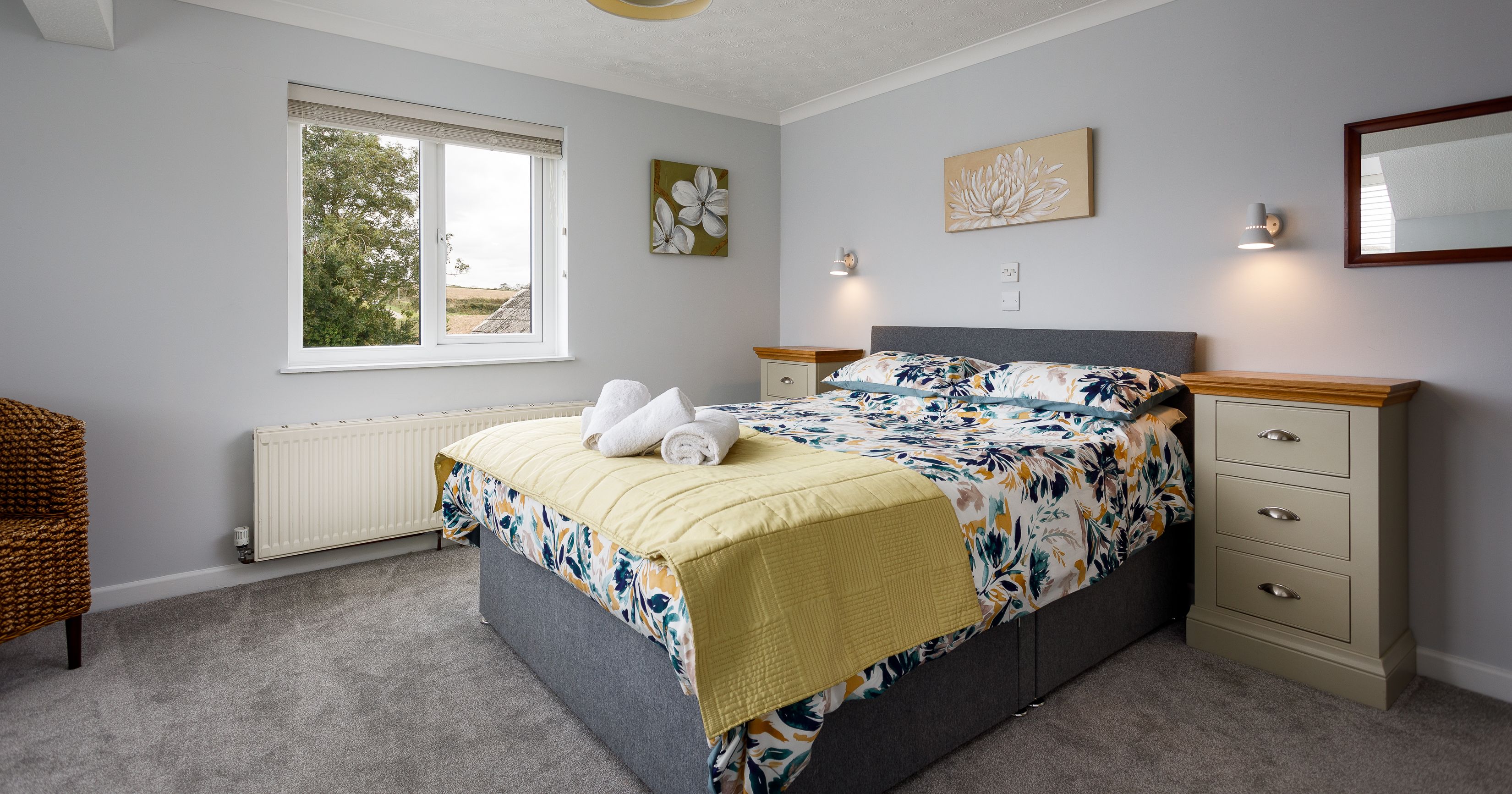 Bright bedroom with a double bed, floral bedding, yellow throw, two bedside tables, modern lighting, window, and decorative wall art.