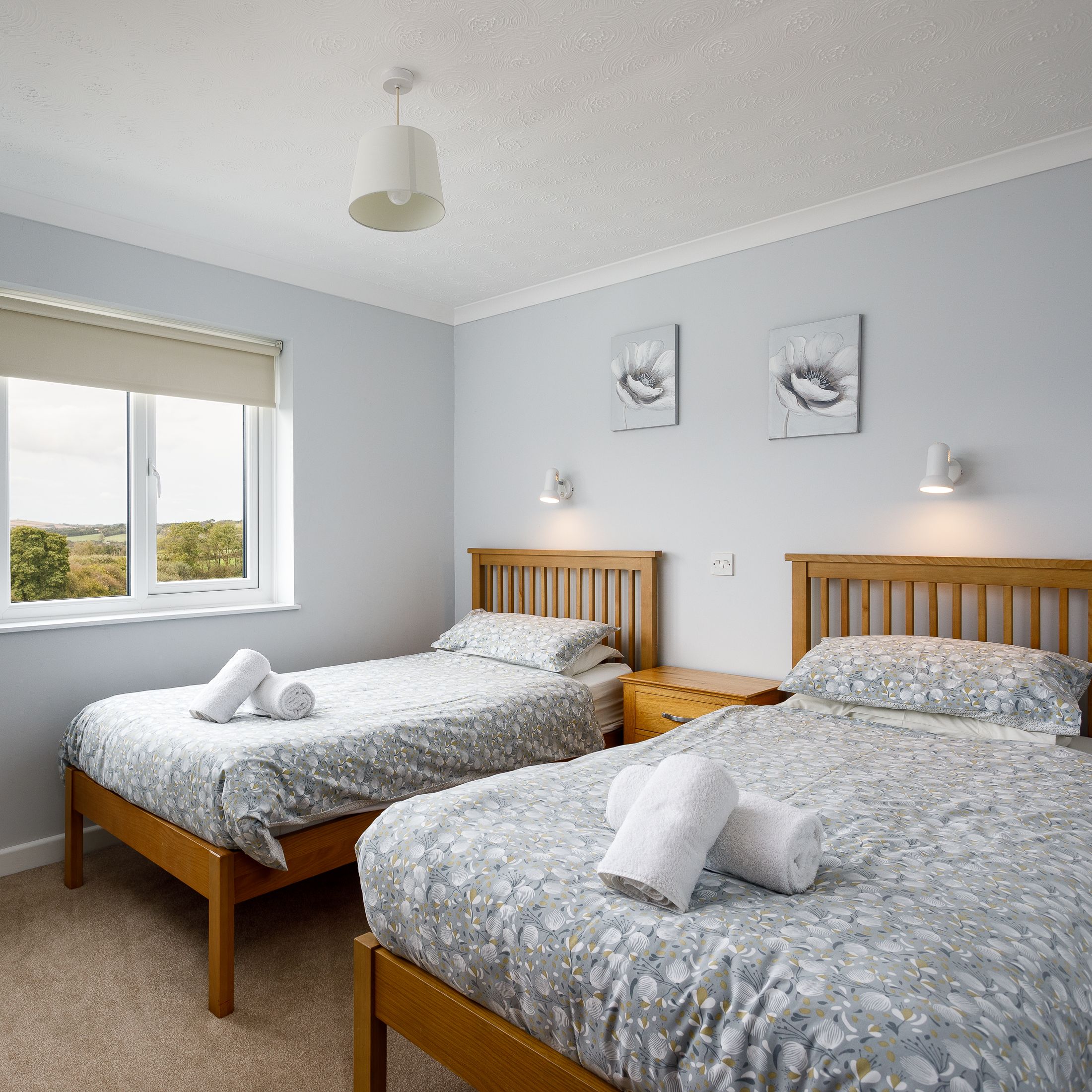 Twin bedroom with wooden beds, grey floral bedding, bedside table, and a large window with countryside view.