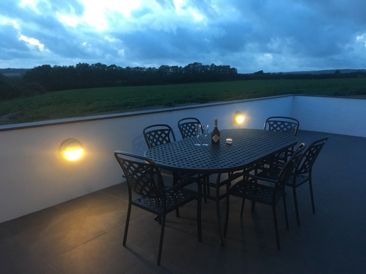 Outdoor patio table with chairs, wine bottle, glasses, and candle at dusk overlooking fields