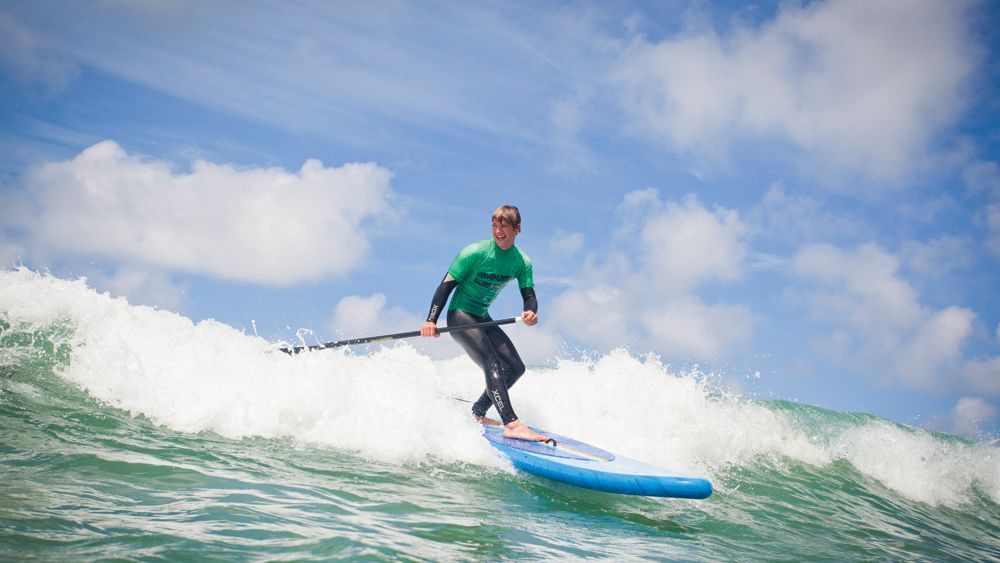 Person paddleboarding on a wave in the ocean under a blue sky with clouds