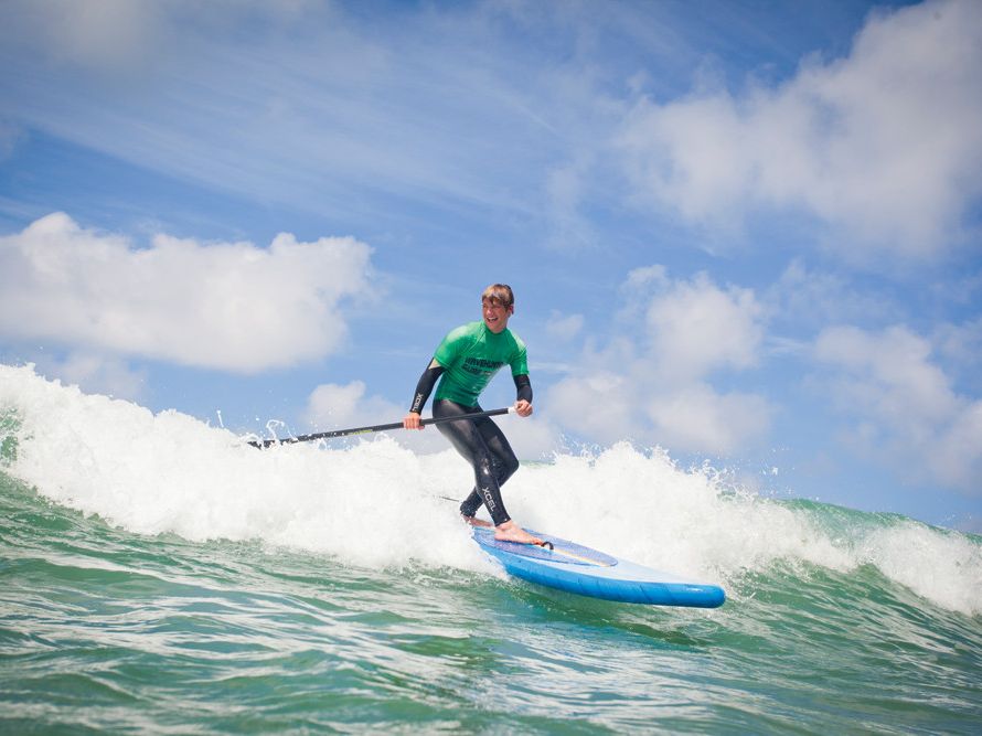 Person paddleboarding on a wave in the ocean under a blue sky with clouds