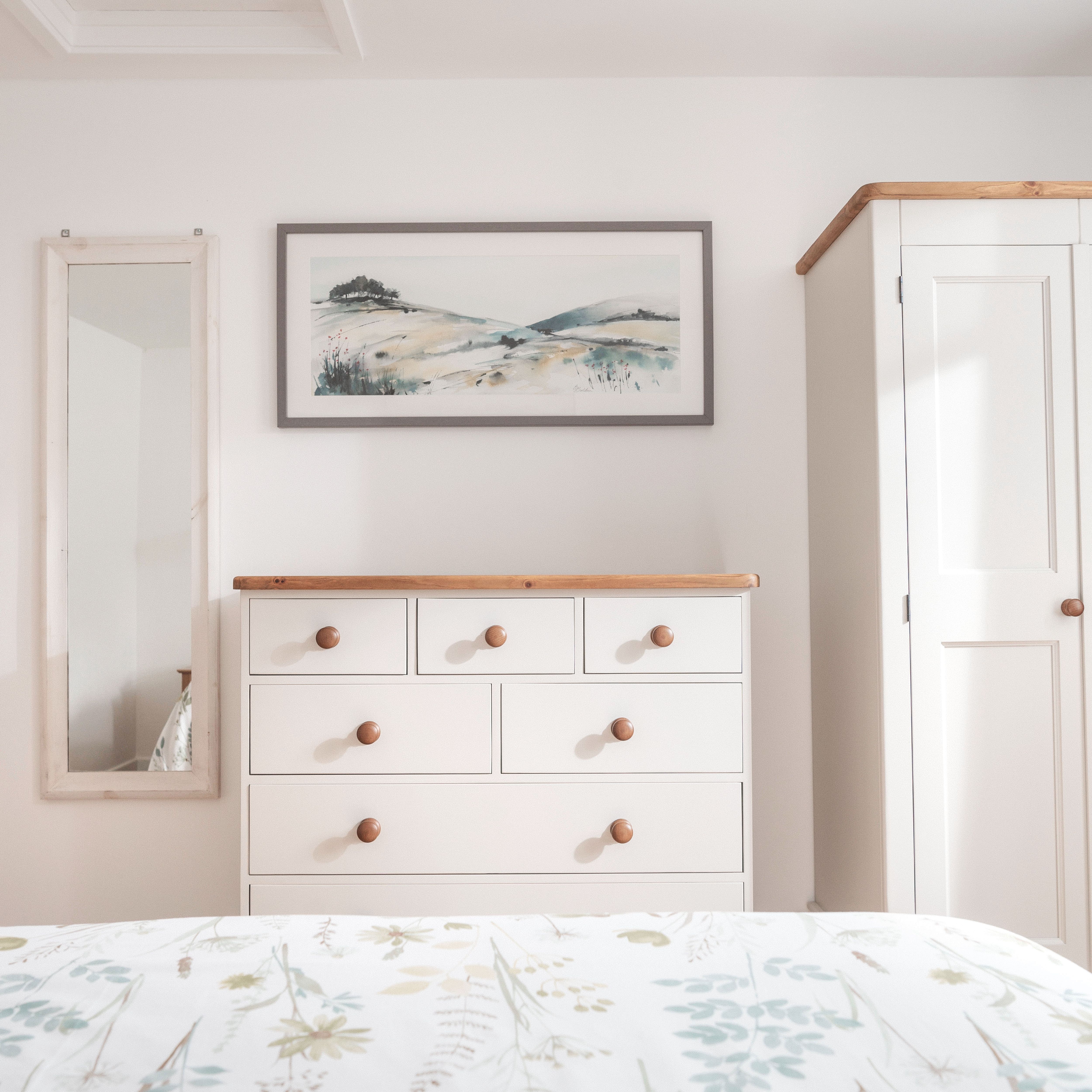 Bright and minimal bedroom with a white dresser, wardrobe, wall mirror, and a landscape painting above the dresser.