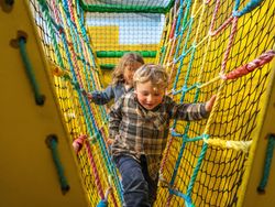 Two young children playing and climbing on a colorful rope bridge in an indoor playground.