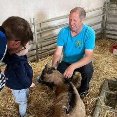 A man and a child interacting with a goat in a straw-covered animal pen.