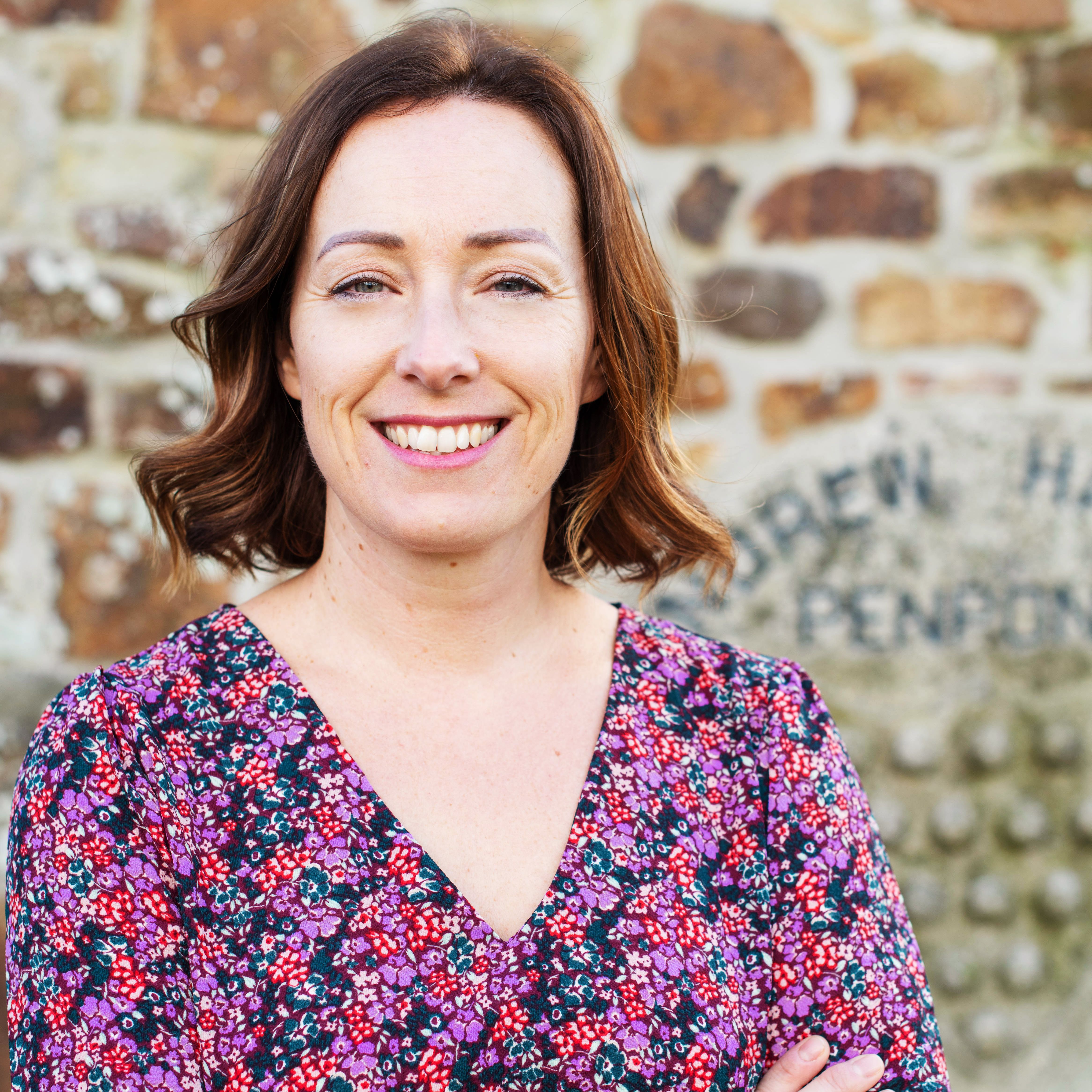 Woman with shoulder-length brown hair wearing a floral dress standing in front of a stone wall