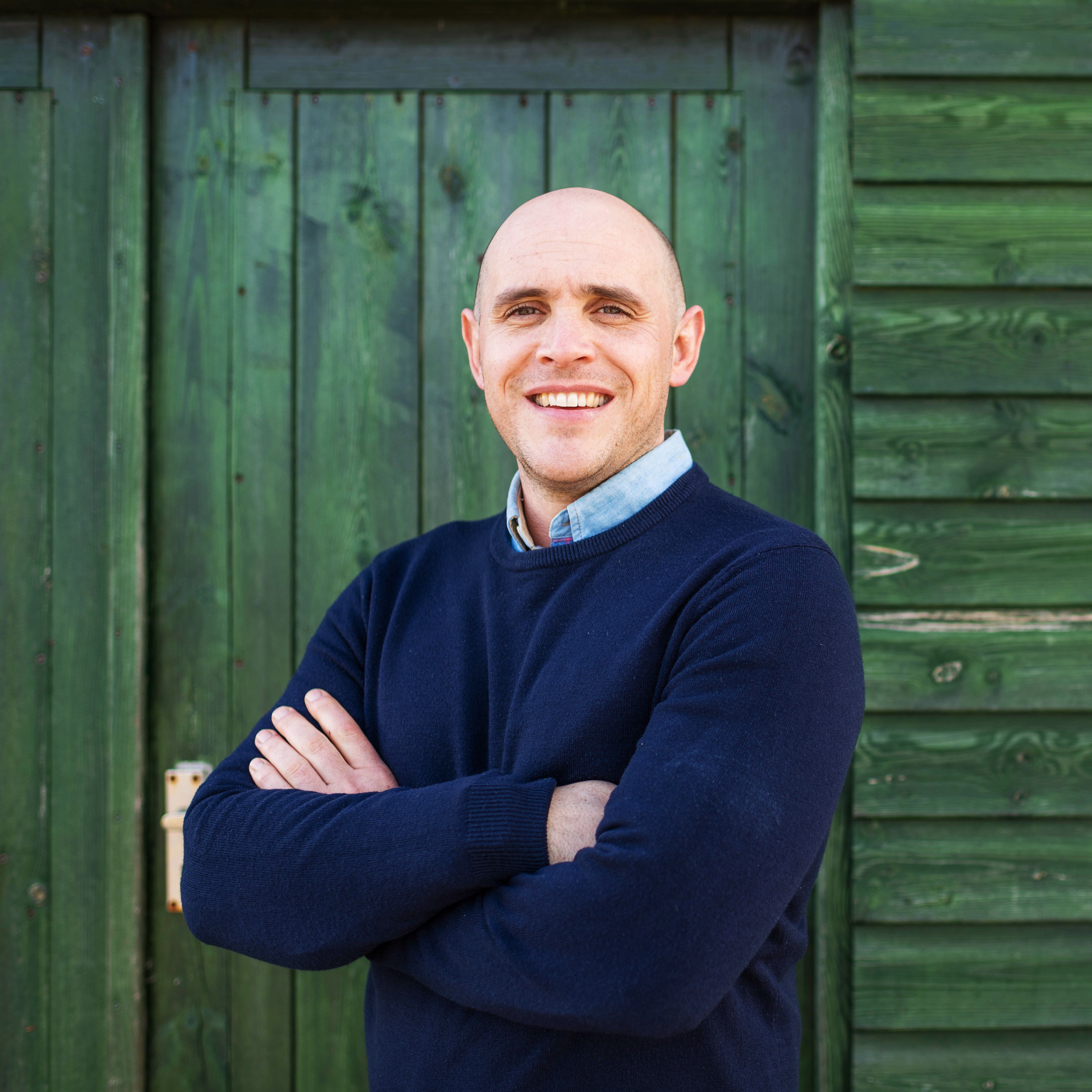 Smiling man with arms crossed standing in front of a green wooden wall