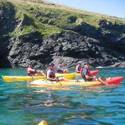 A group of people kayaking on clear turquoise water near rocky cliffs with green grass under a bright blue sky.