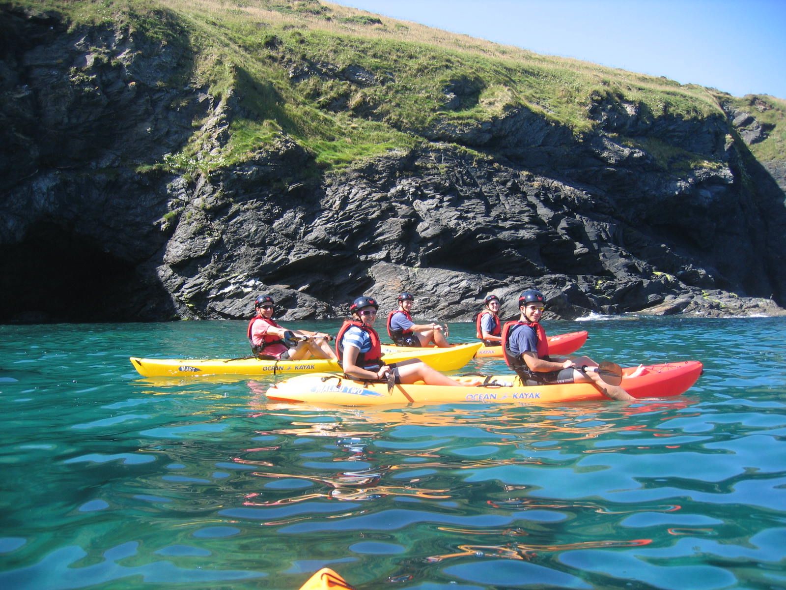 A group of people kayaking on clear turquoise water near rocky cliffs with green grass under a bright blue sky.