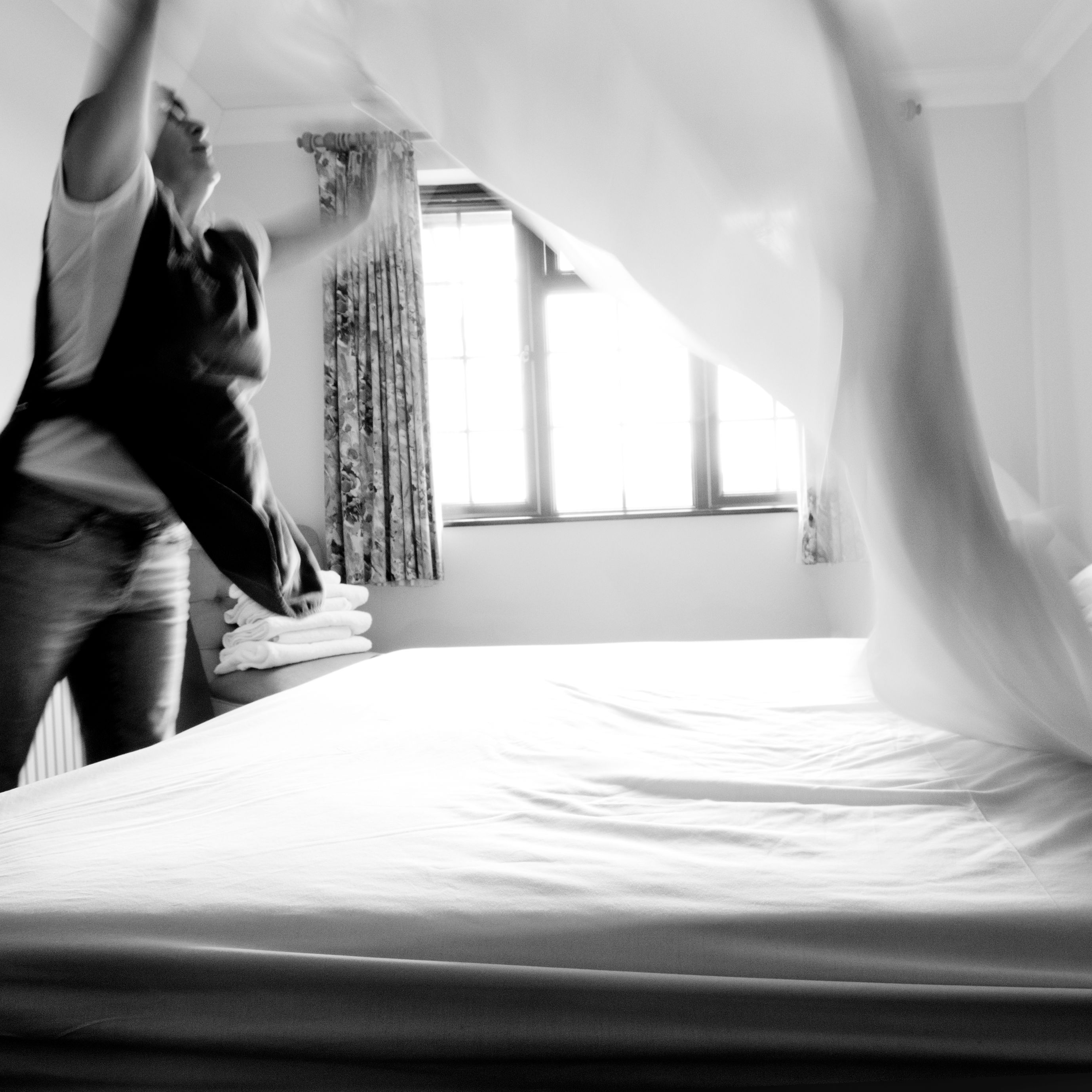 Person making a bed in a bright room, throwing a sheet onto the mattress.