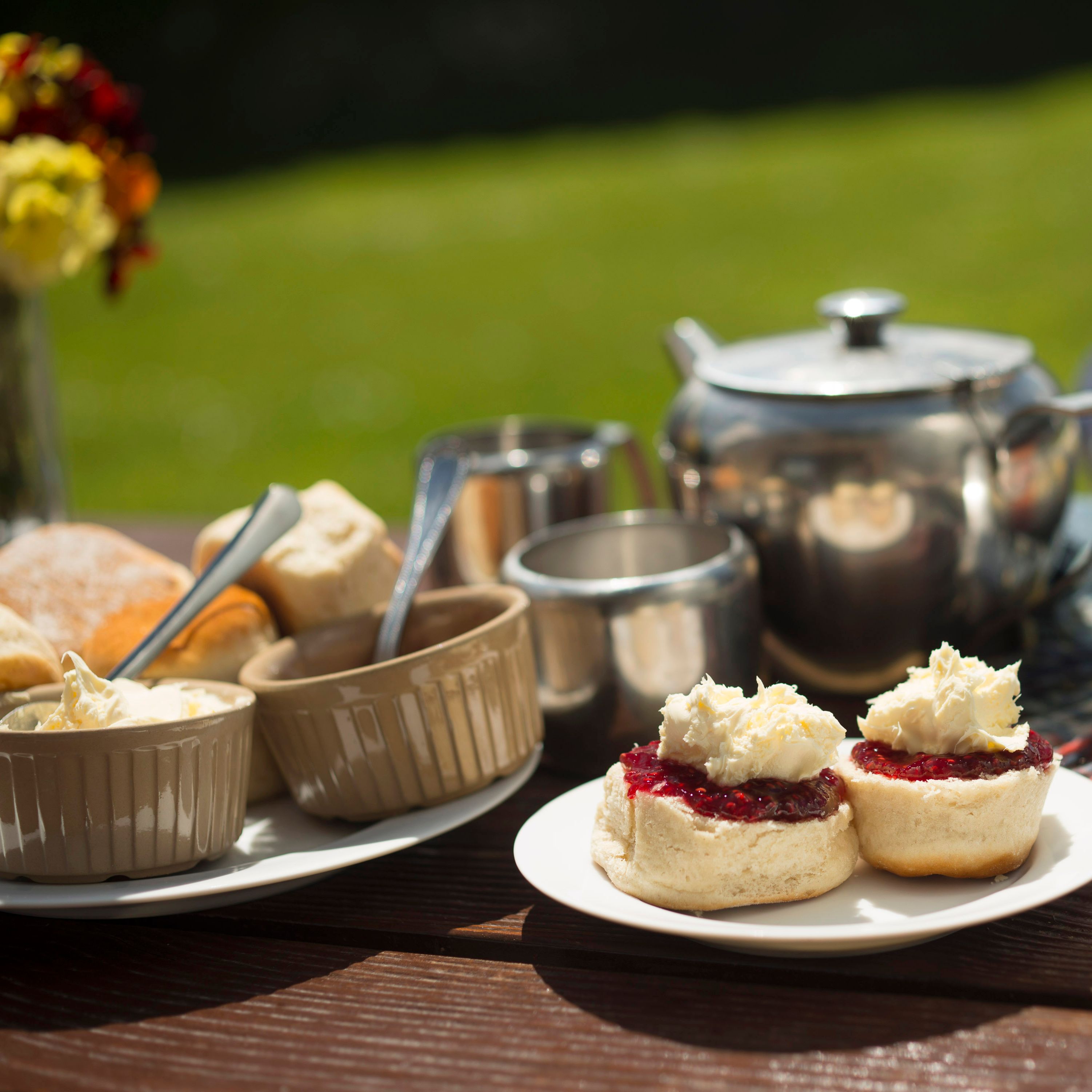 Afternoon cream tea setup with scones, jam, clotted cream, and a teapot on a picnic table outdoors.