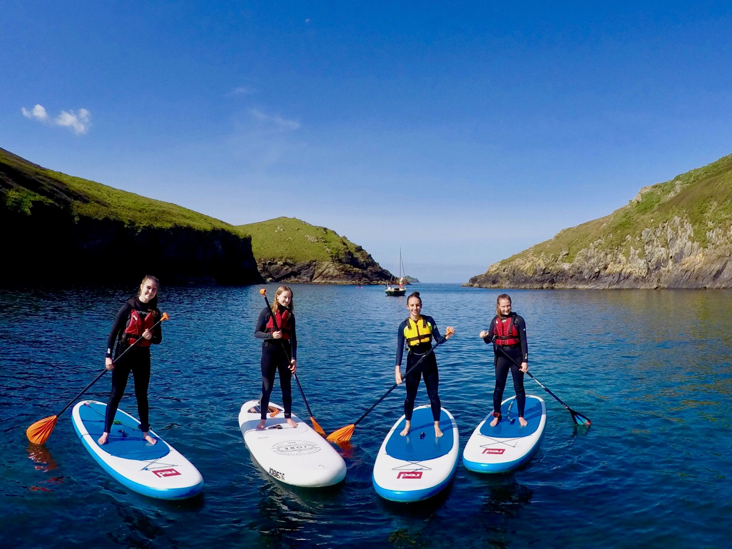 Four people stand on paddleboards in a scenic coastal inlet, wearing wetsuits and life jackets.