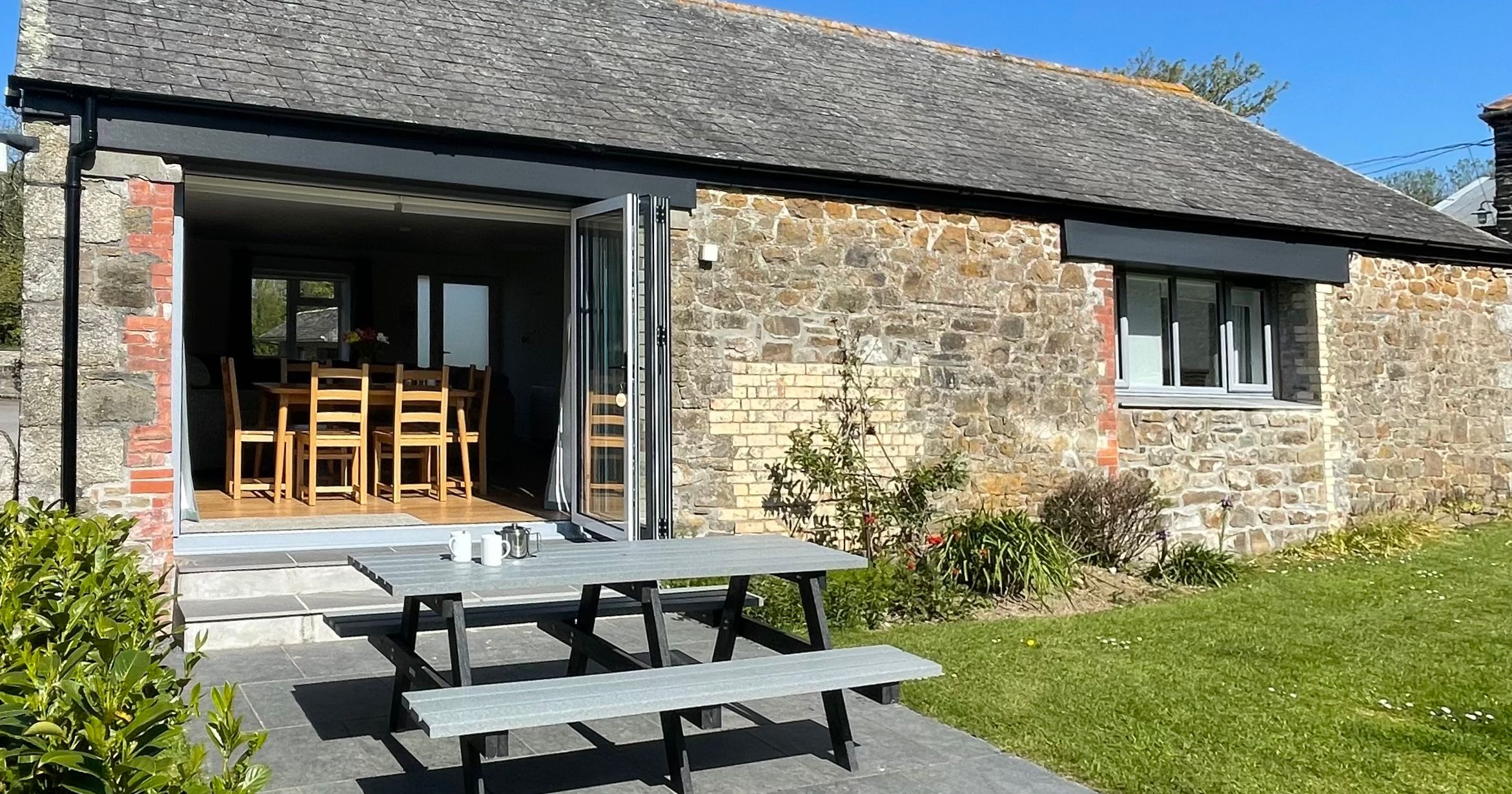 Stone barn with open doors, outdoor picnic table, and grassy garden area under a clear blue sky.