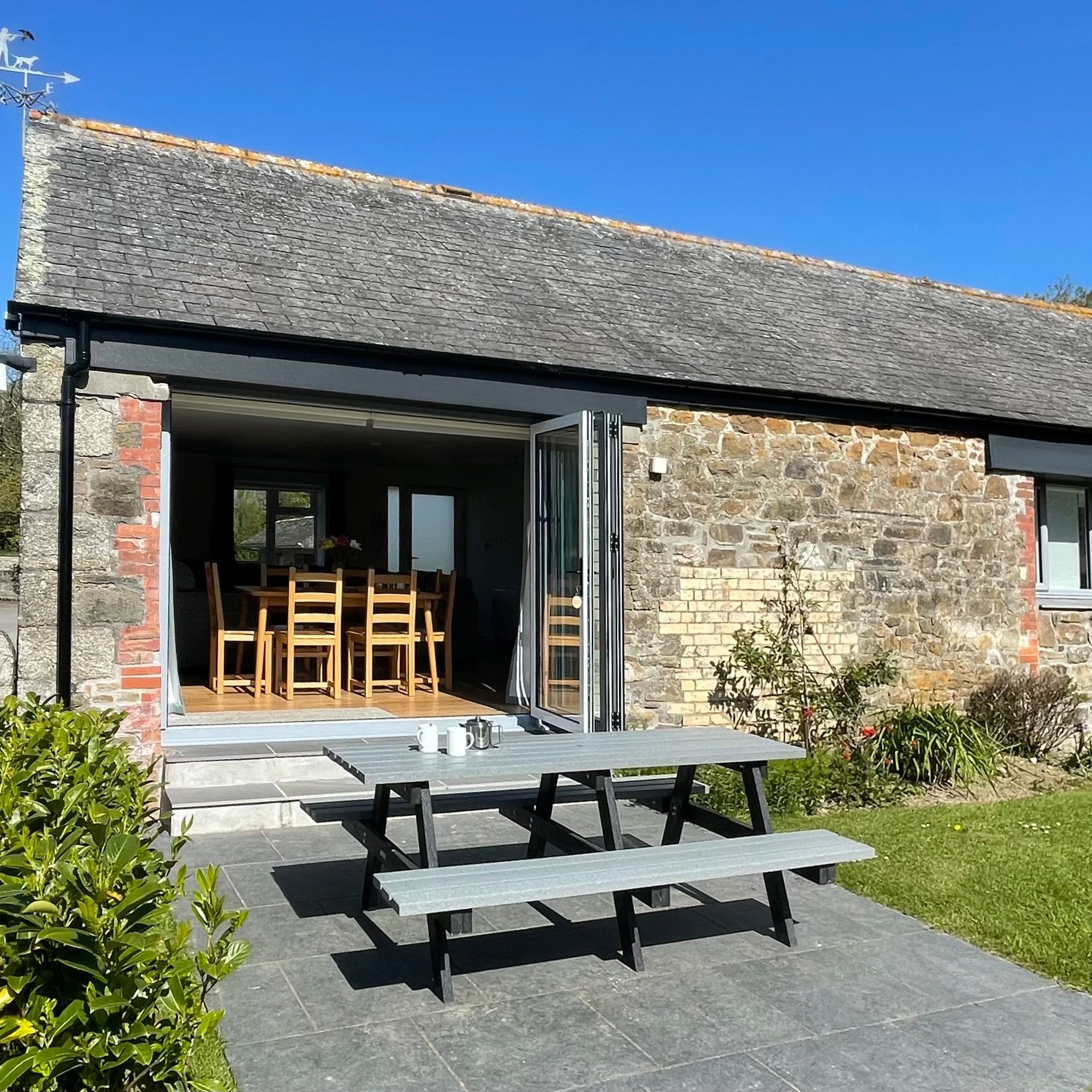 Stone barn with open doors, outdoor picnic table, and grassy garden area under a clear blue sky.