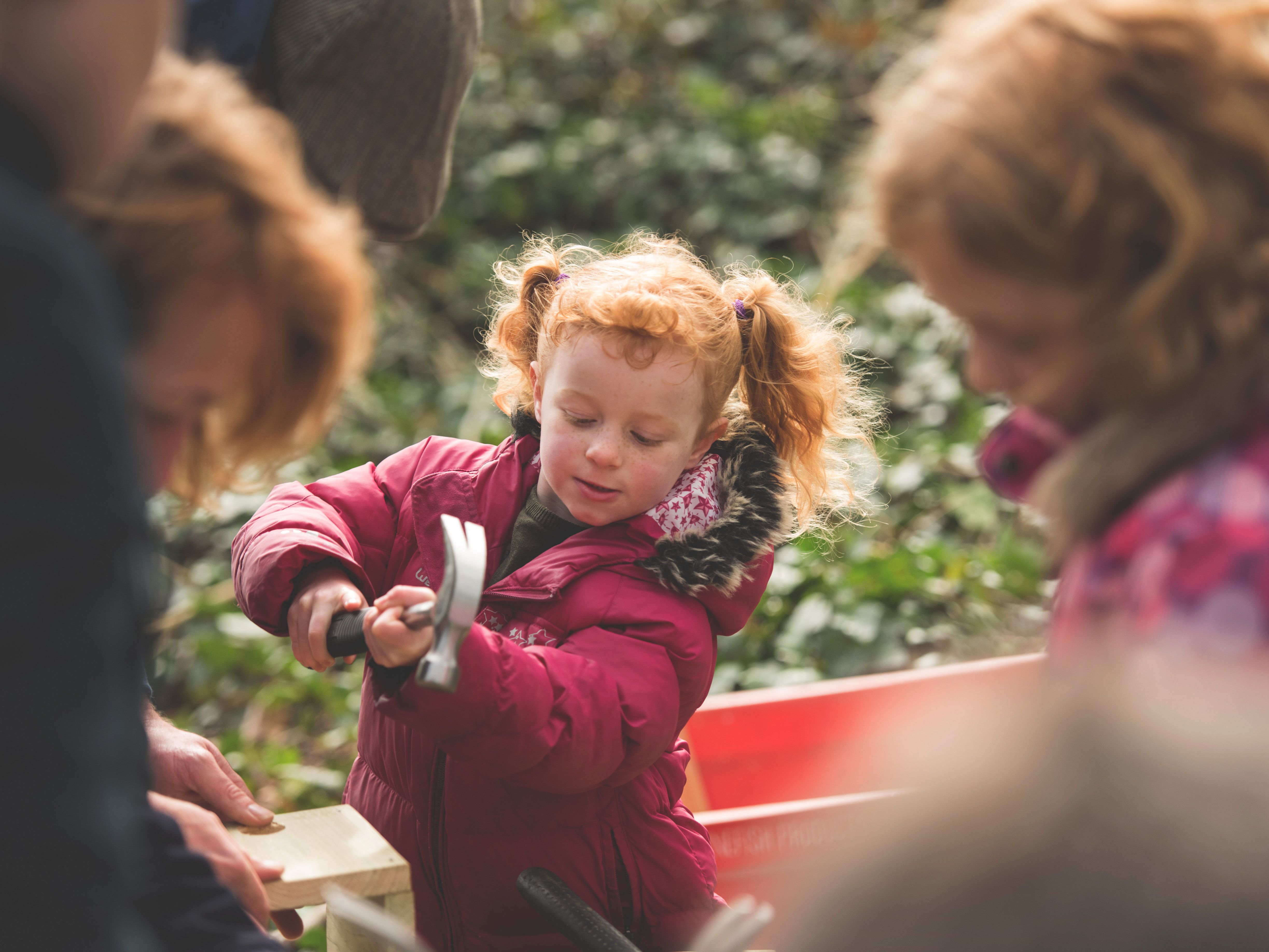 Young girl with red hair in pigtails wearing a maroon jacket, using a hammer outdoors with other children and adults.