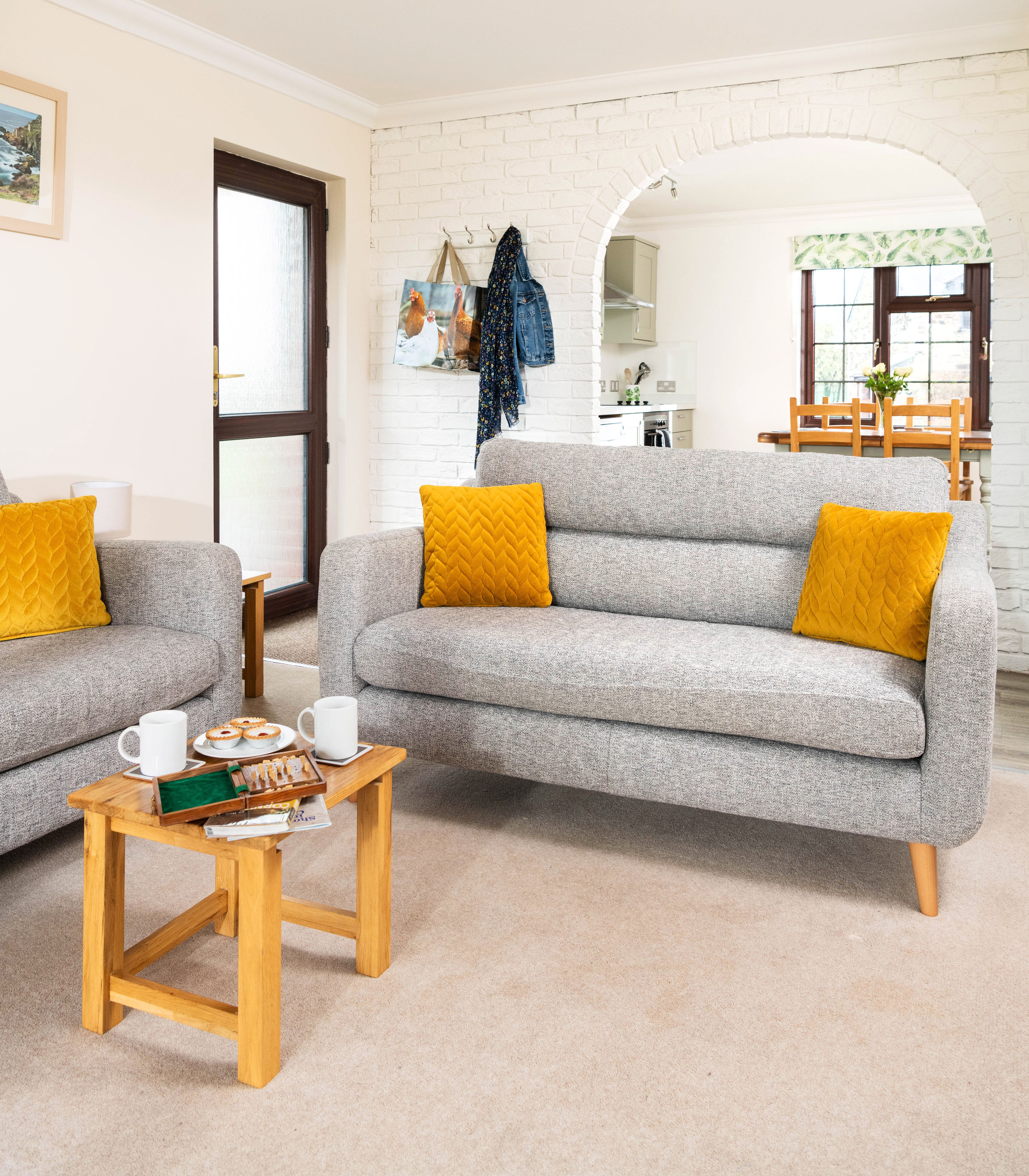 Cozy living room with grey sofas, yellow cushions, a wooden coffee table with tea and treats, and a view into a bright kitchen through a white brick archway.
