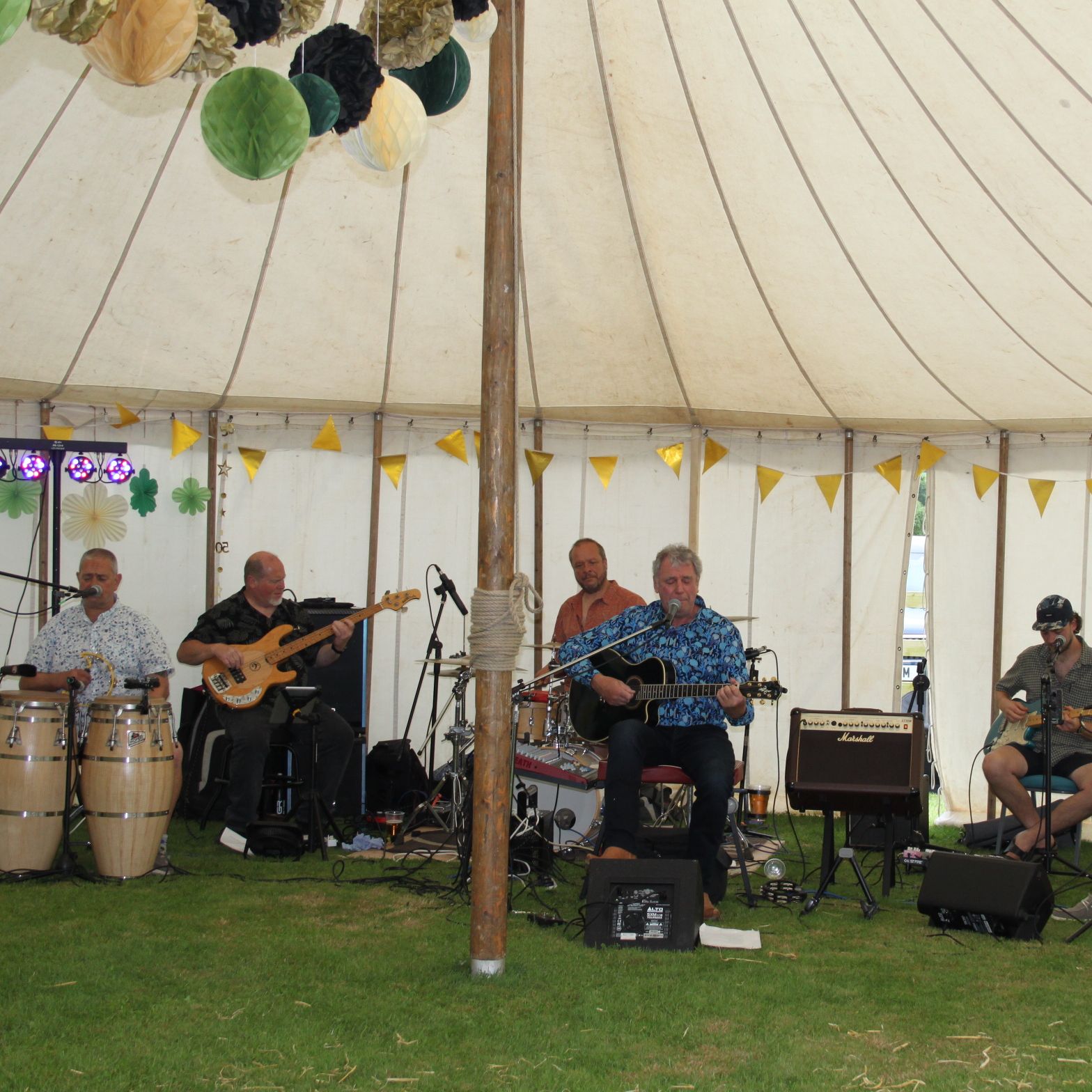 A band of six musicians playing various instruments inside a large white tent decorated with bunting and paper decorations.