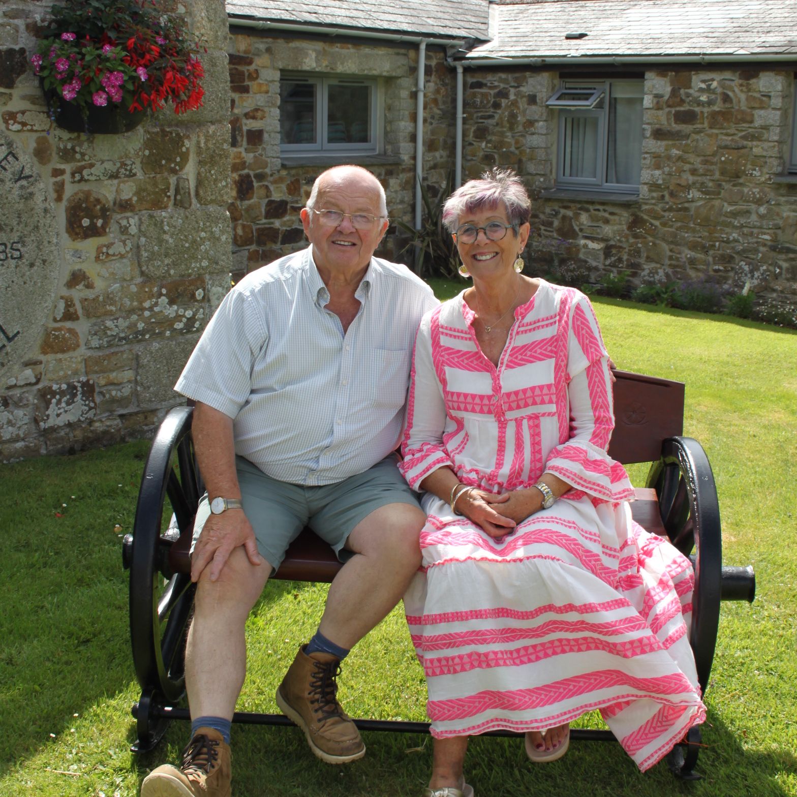 Older man and woman sitting together on a bench outside a stone building on a sunny day