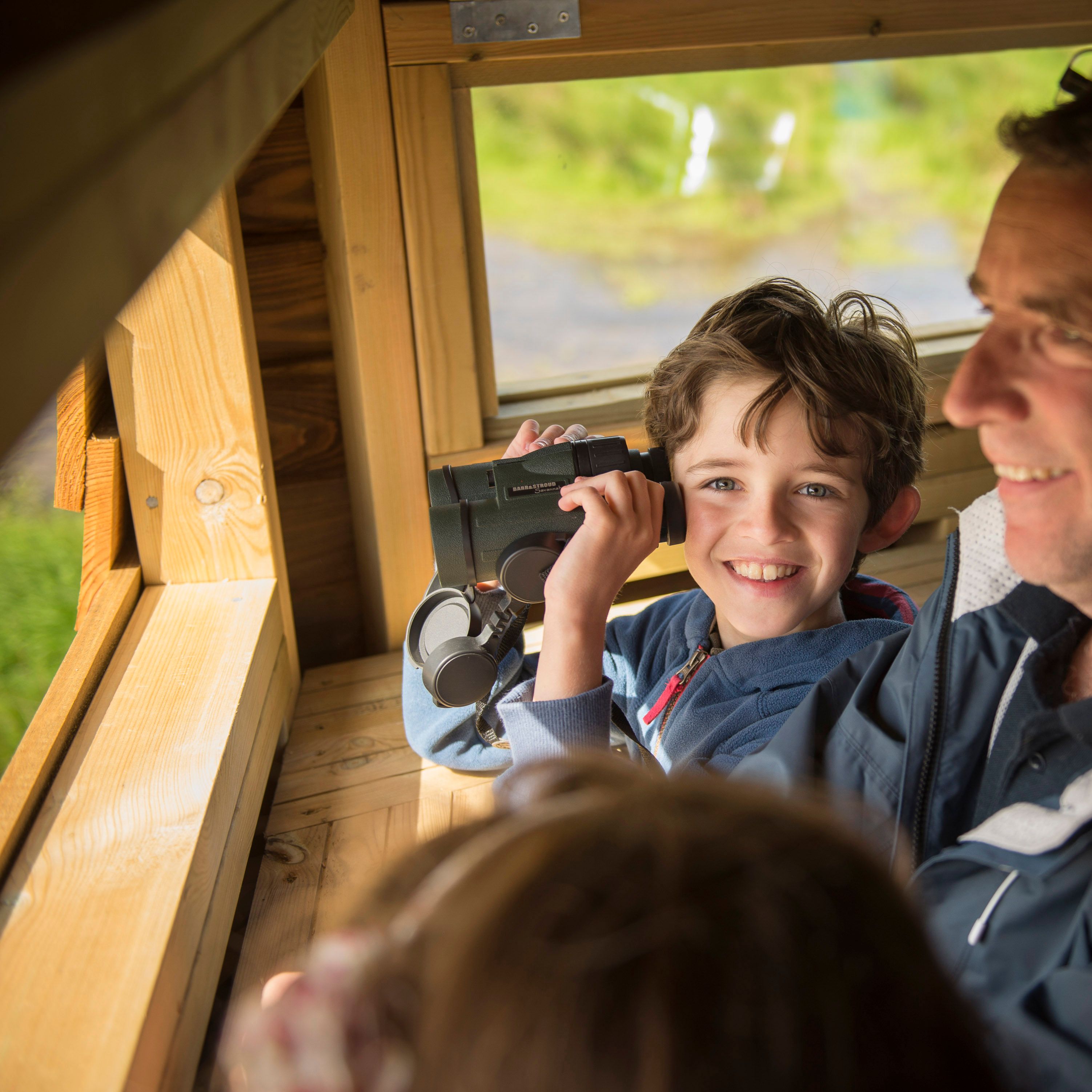 A smiling boy with binoculars sits beside an adult man inside a wooden bird hide, looking out the window at nature.