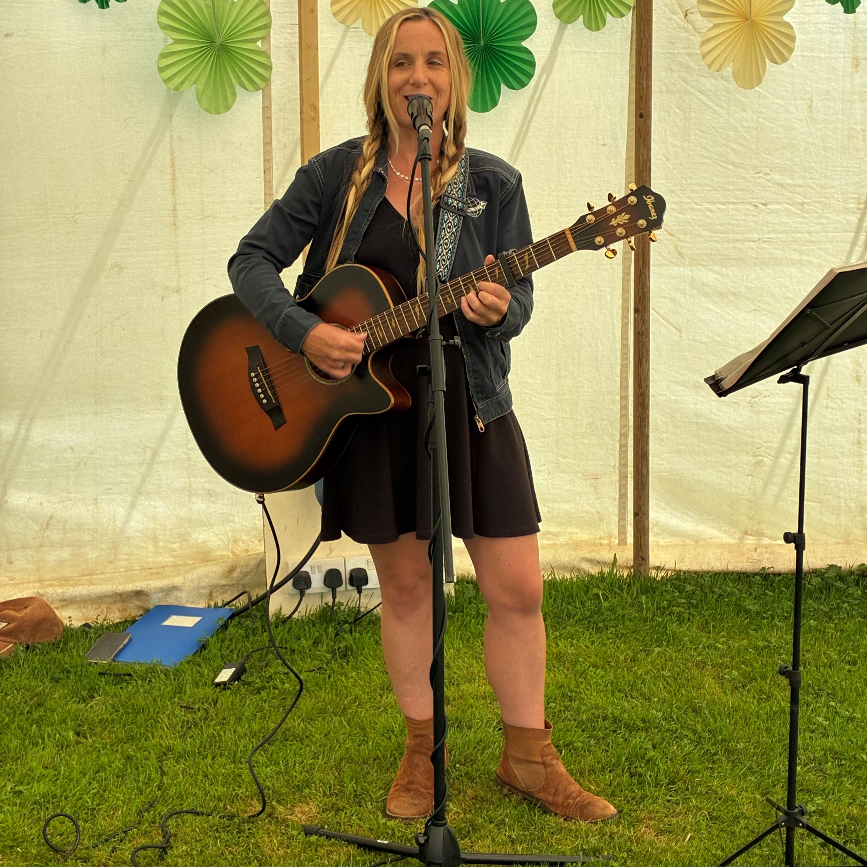 Woman playing acoustic guitar and singing into a microphone at an outdoor event tent decorated with green and yellow paper flowers.