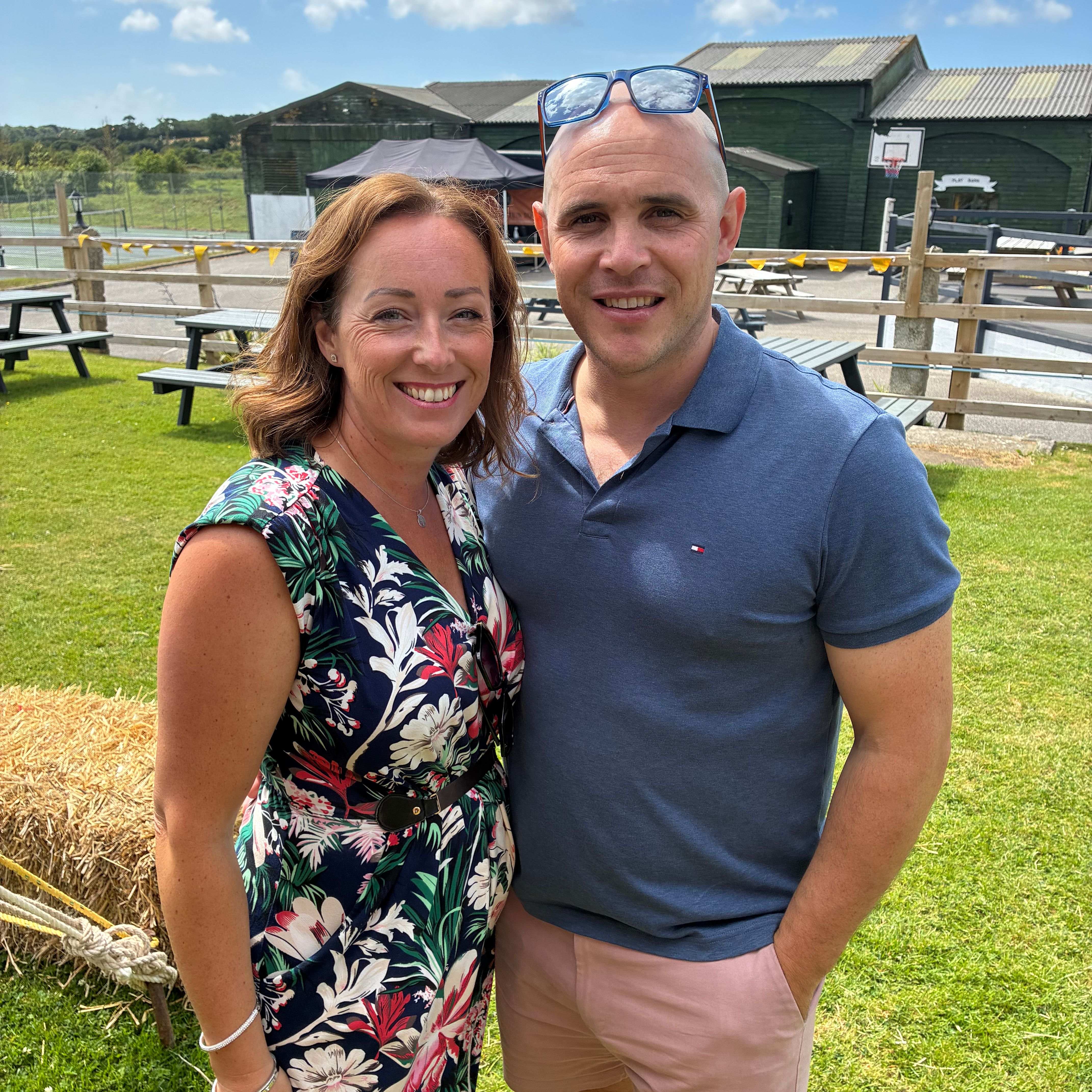 A smiling couple posing together outdoors on a sunny day, standing on grass near hay bales and picnic benches.