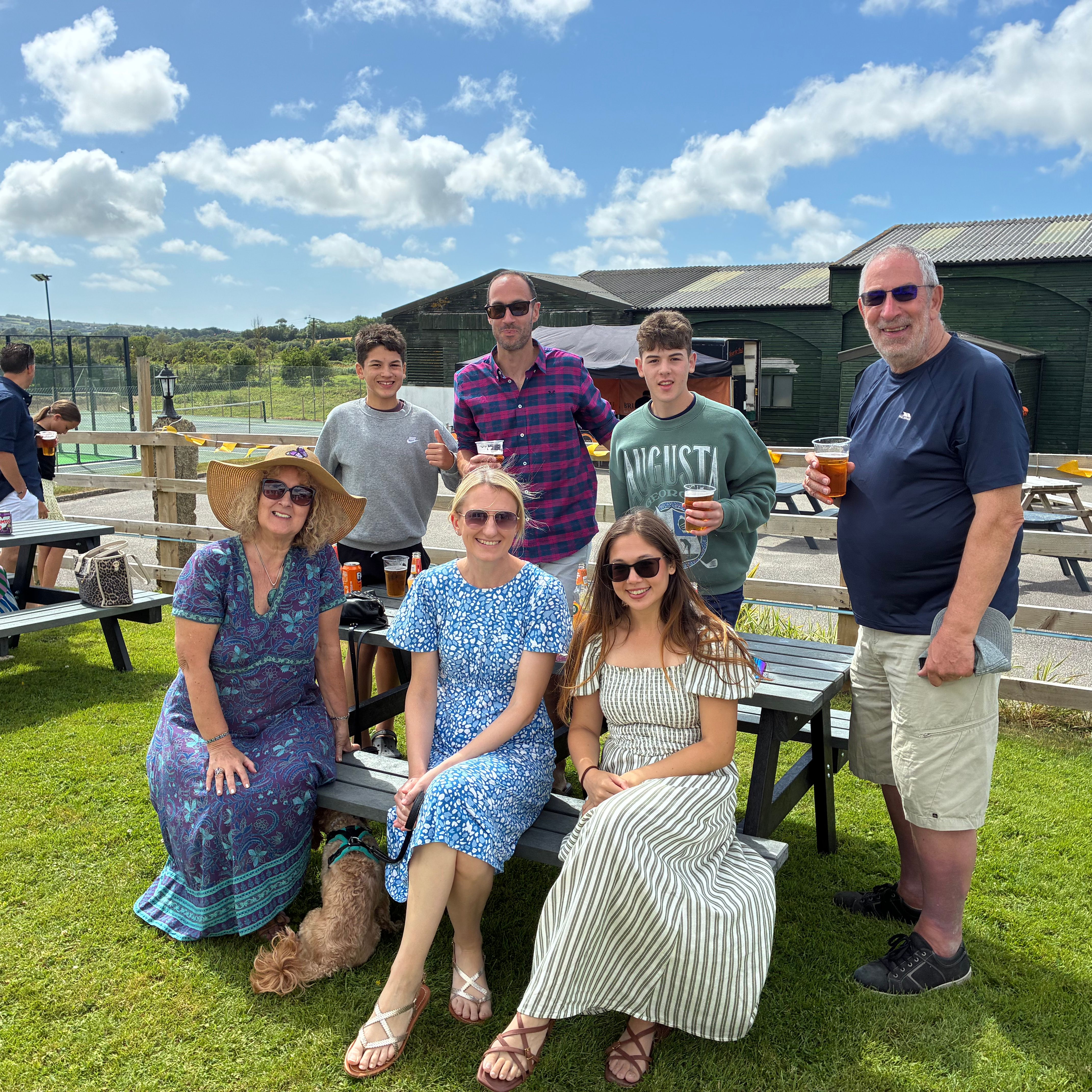 Group of people enjoying a sunny outdoor gathering near picnic benches with drinks, tennis courts in the background.