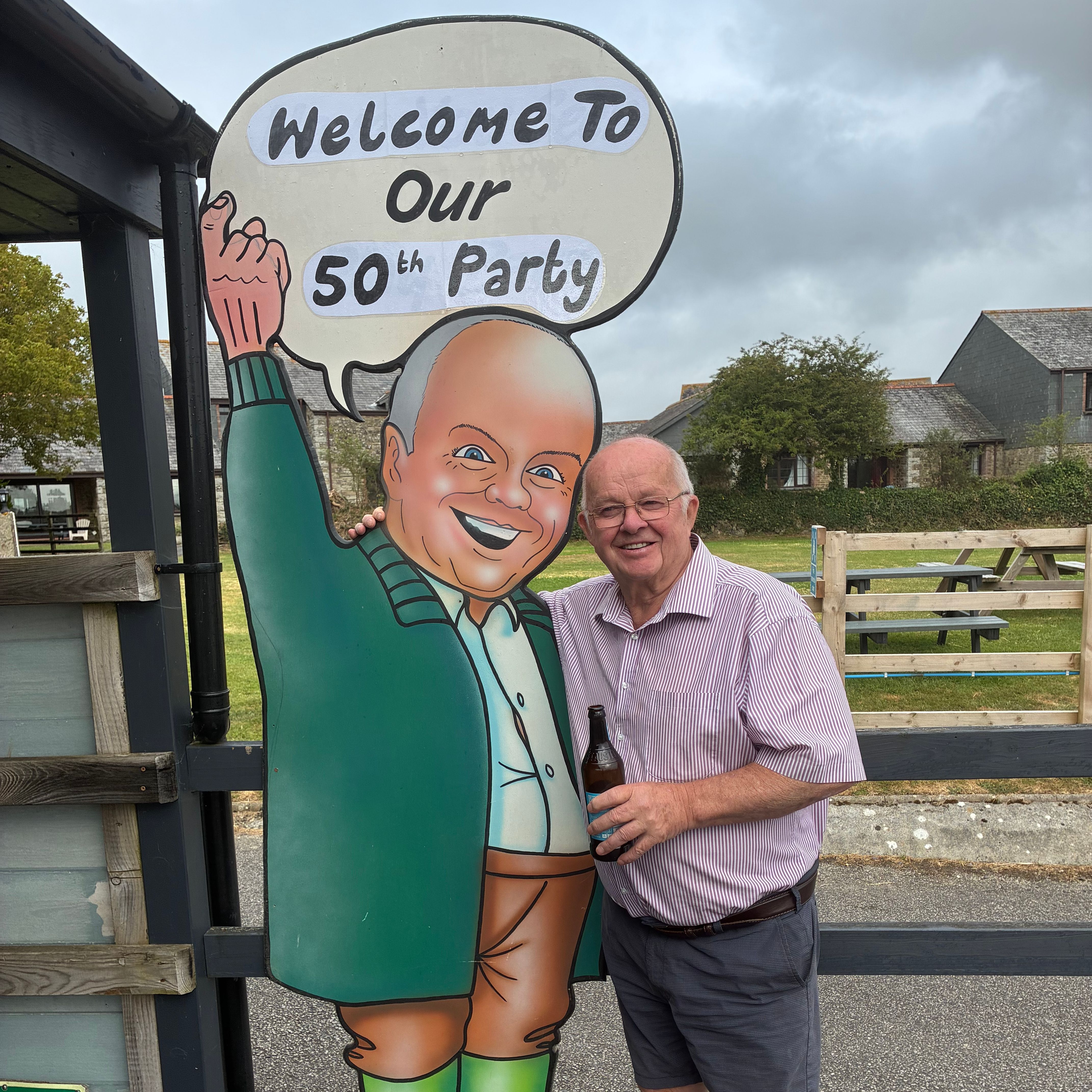 Man standing next to a large cartoon cutout with a speech bubble that says 'Welcome To Our 50th Party'.