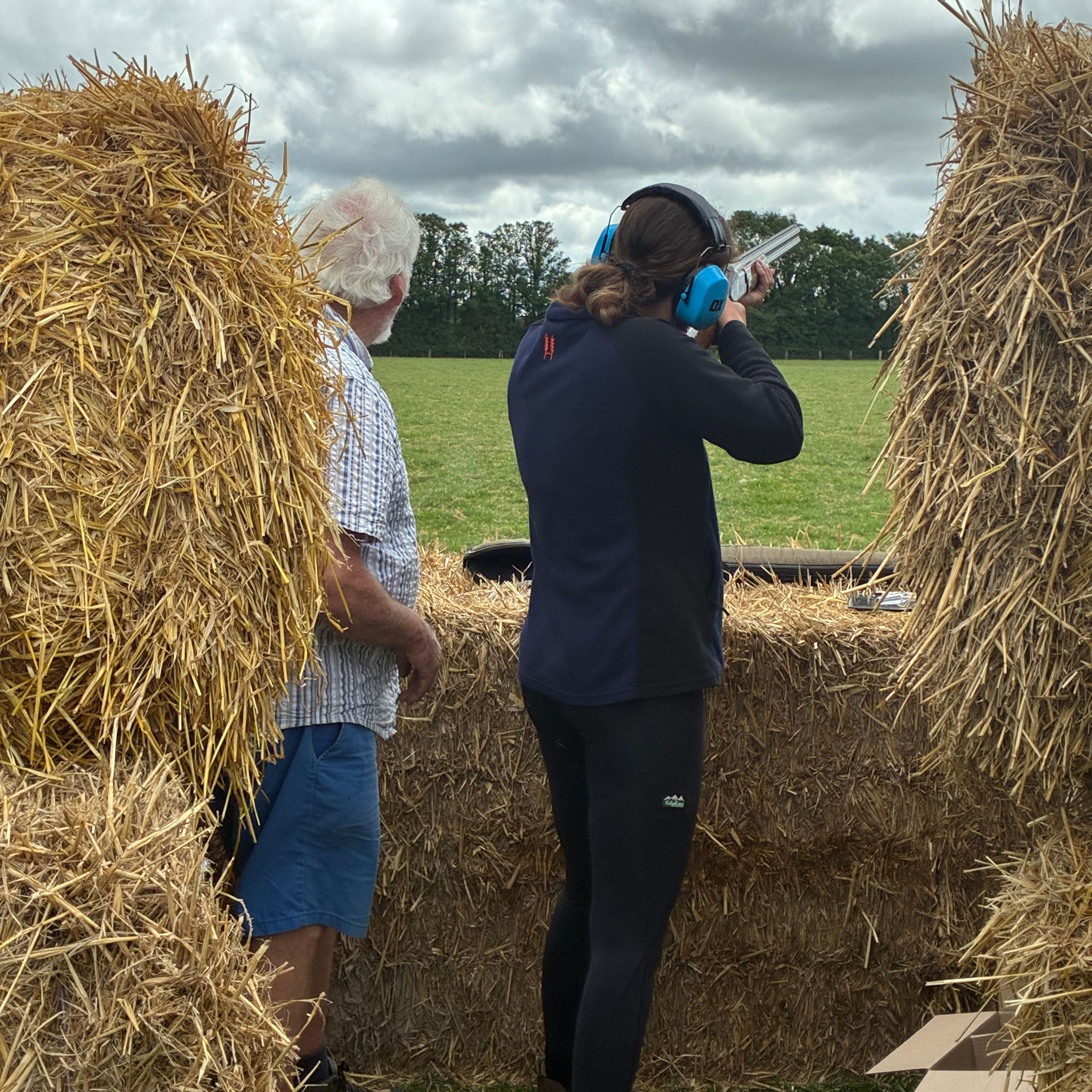 Person aiming a shotgun at an outdoor shooting range with hay bales and another person standing beside them.