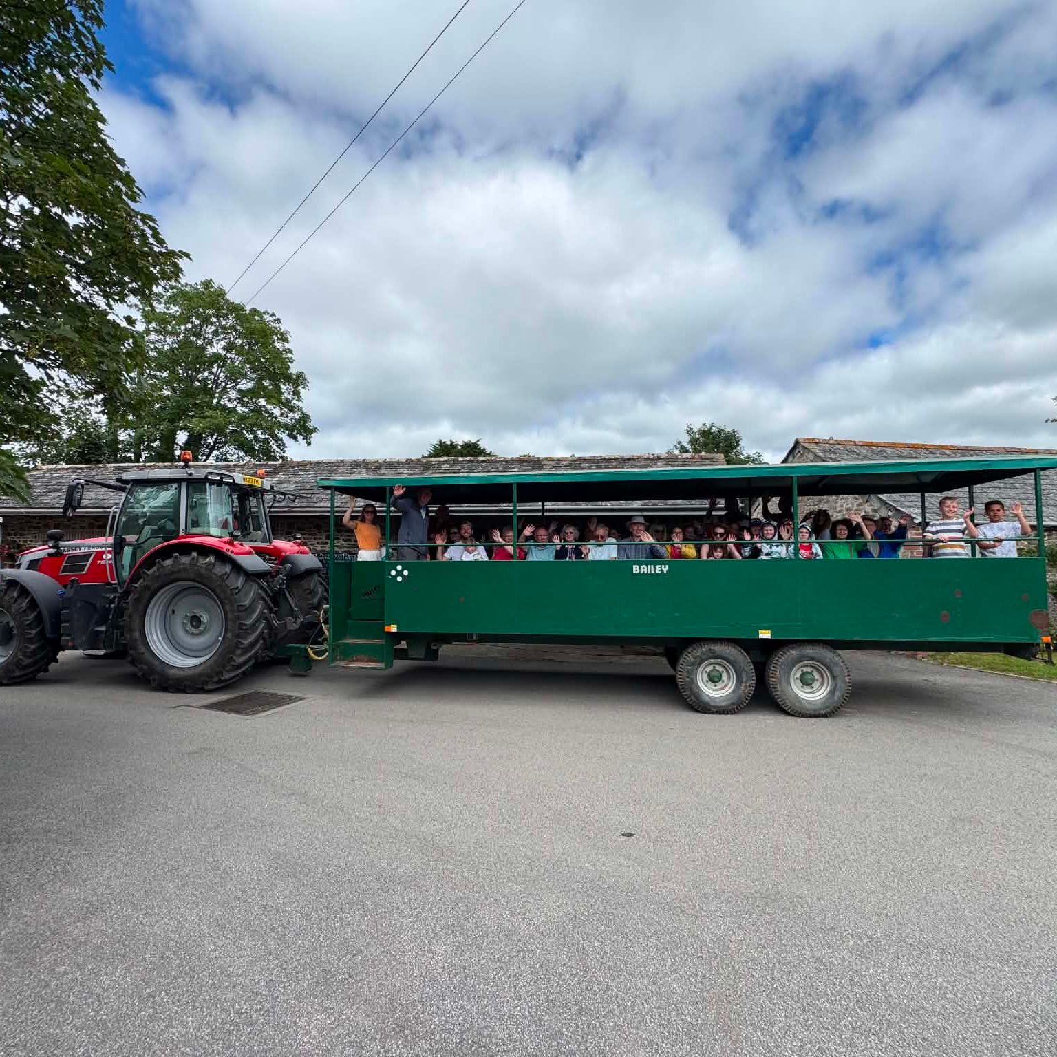 A red tractor pulling a green trailer filled with people, passing by rustic buildings under a partly cloudy sky.