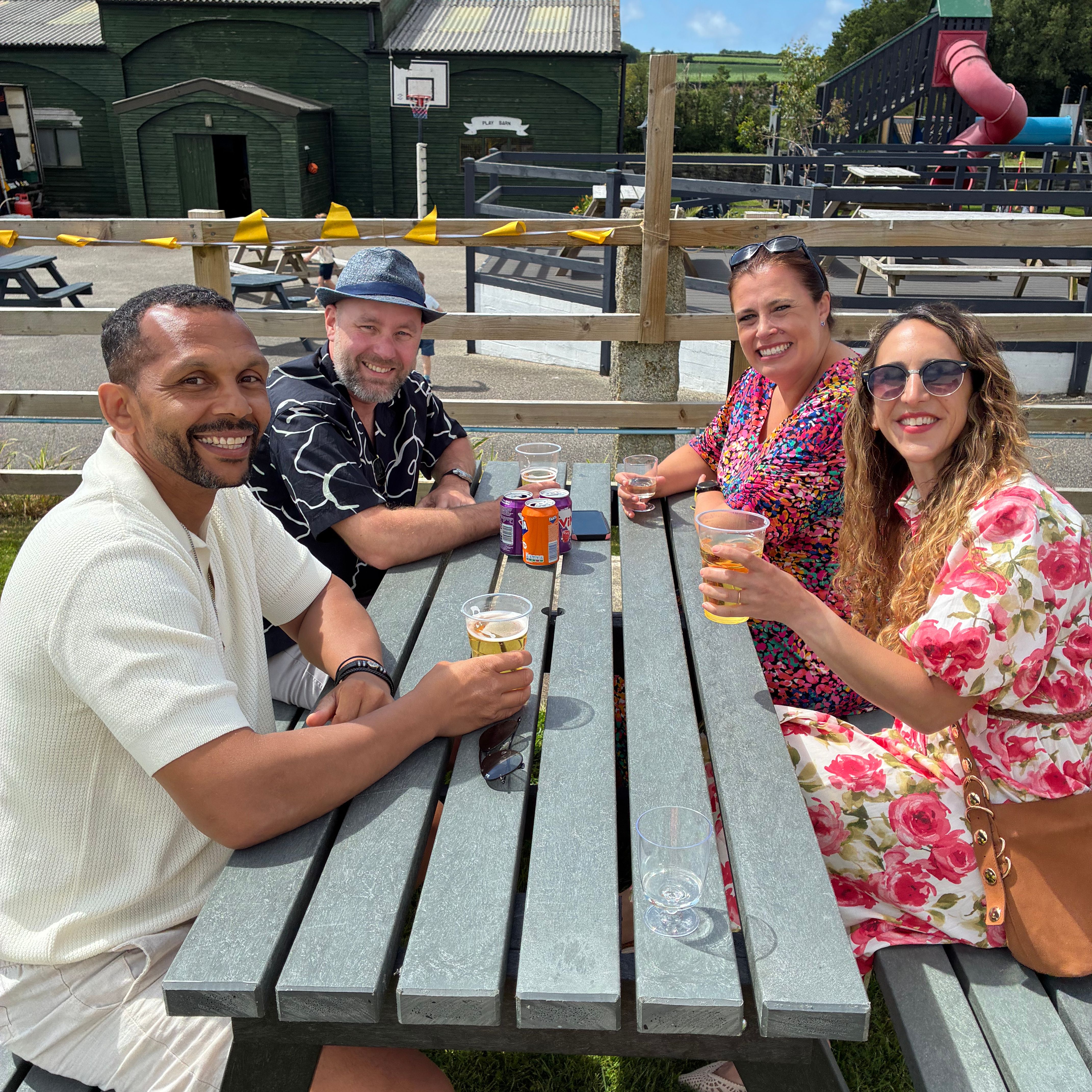 Four adults sitting at an outdoor picnic table, smiling and enjoying drinks on a sunny day.