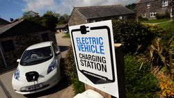 Electric vehicle charging station sign in front of a white electric car that is plugged in, with stone buildings and greenery in the background.