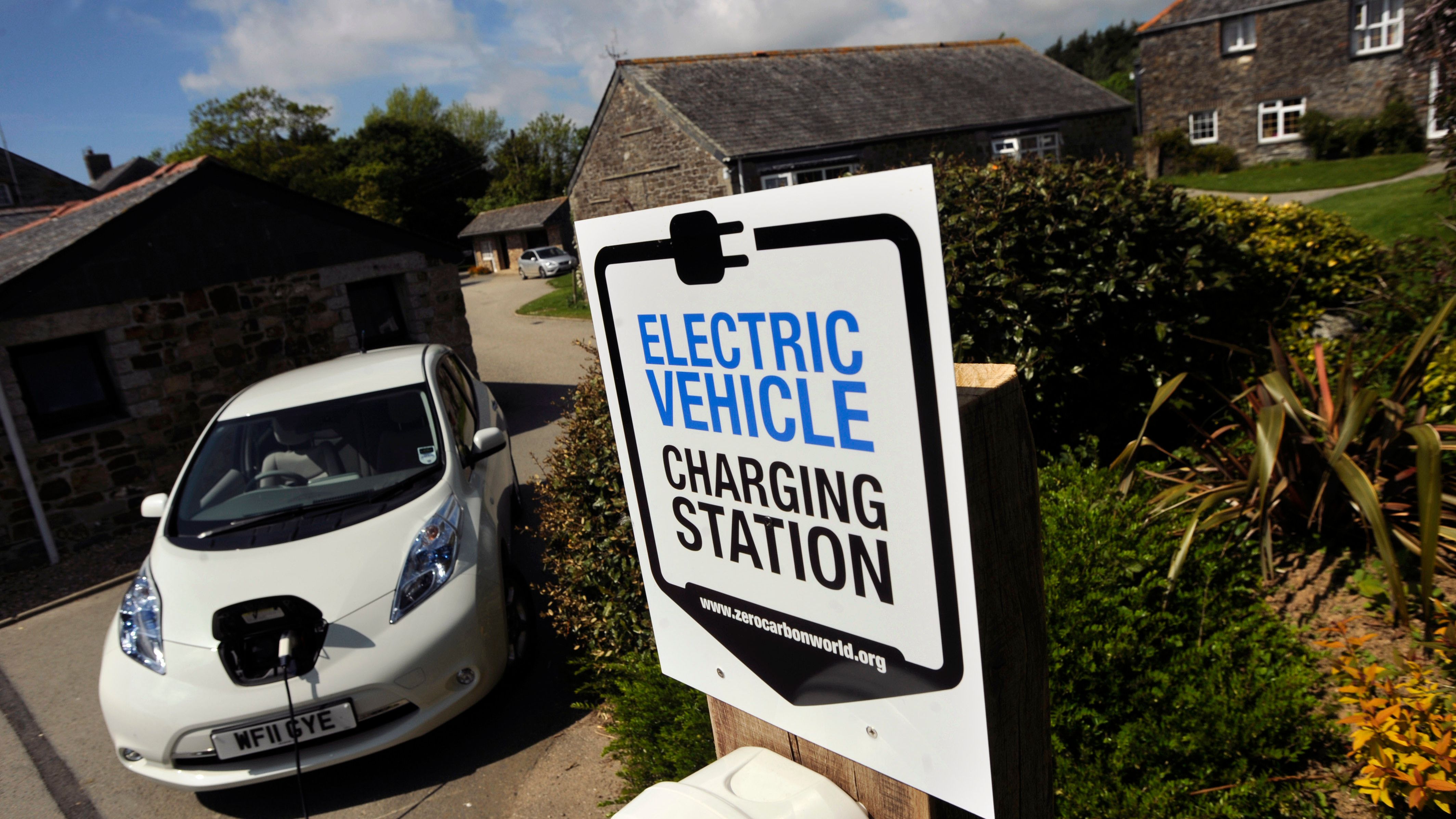Electric vehicle charging station sign in front of a white electric car that is plugged in, with stone buildings and greenery in the background.